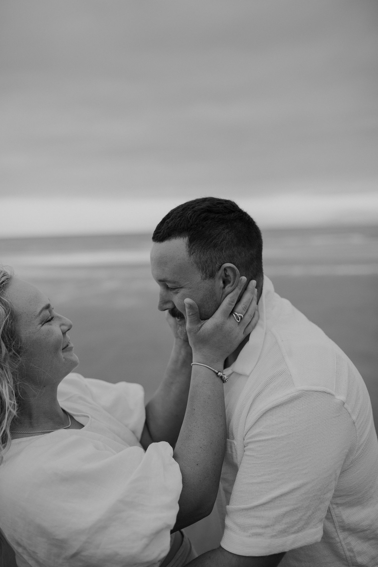 Couple engagement photo session. Black and white, holding mans face and smiling. Beach at Kina.