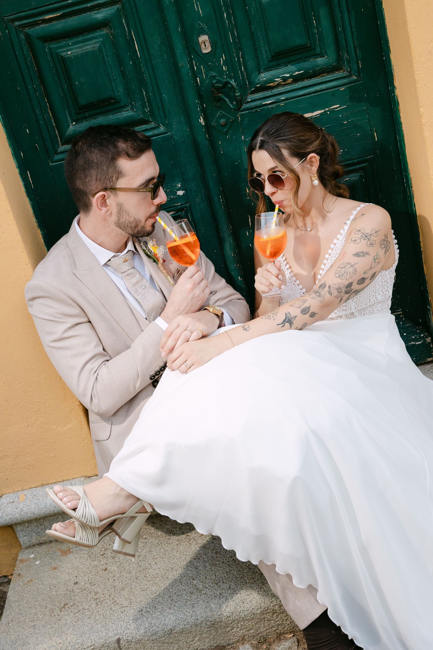 Wedding couple in sunglasses sipping Aperol Spritz on steps before a green door, bride in white gown, groom in beige suit.