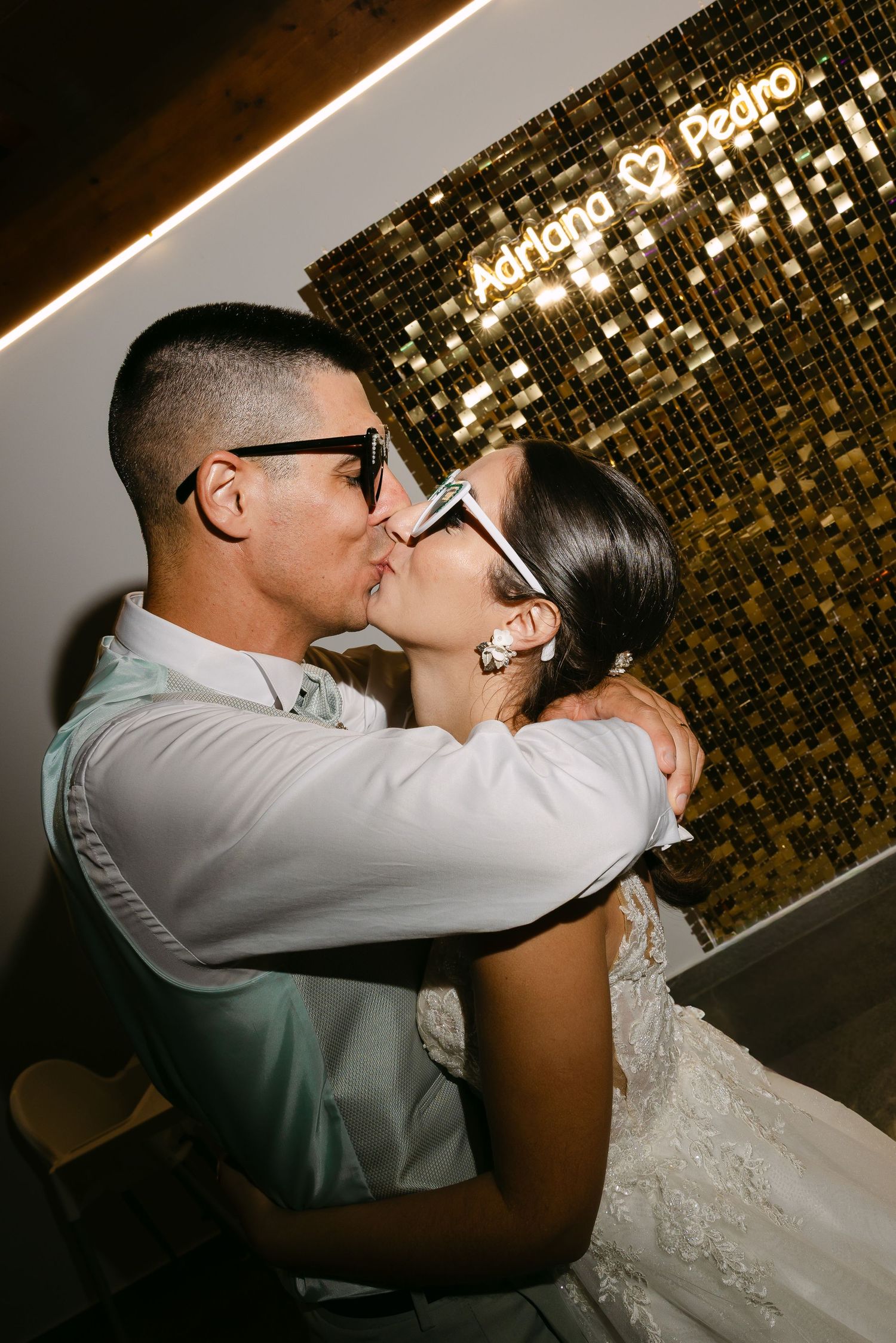 Bride and groom kissing in front of a gold sequin wall with a neon sign reading Adriana and Pedro.