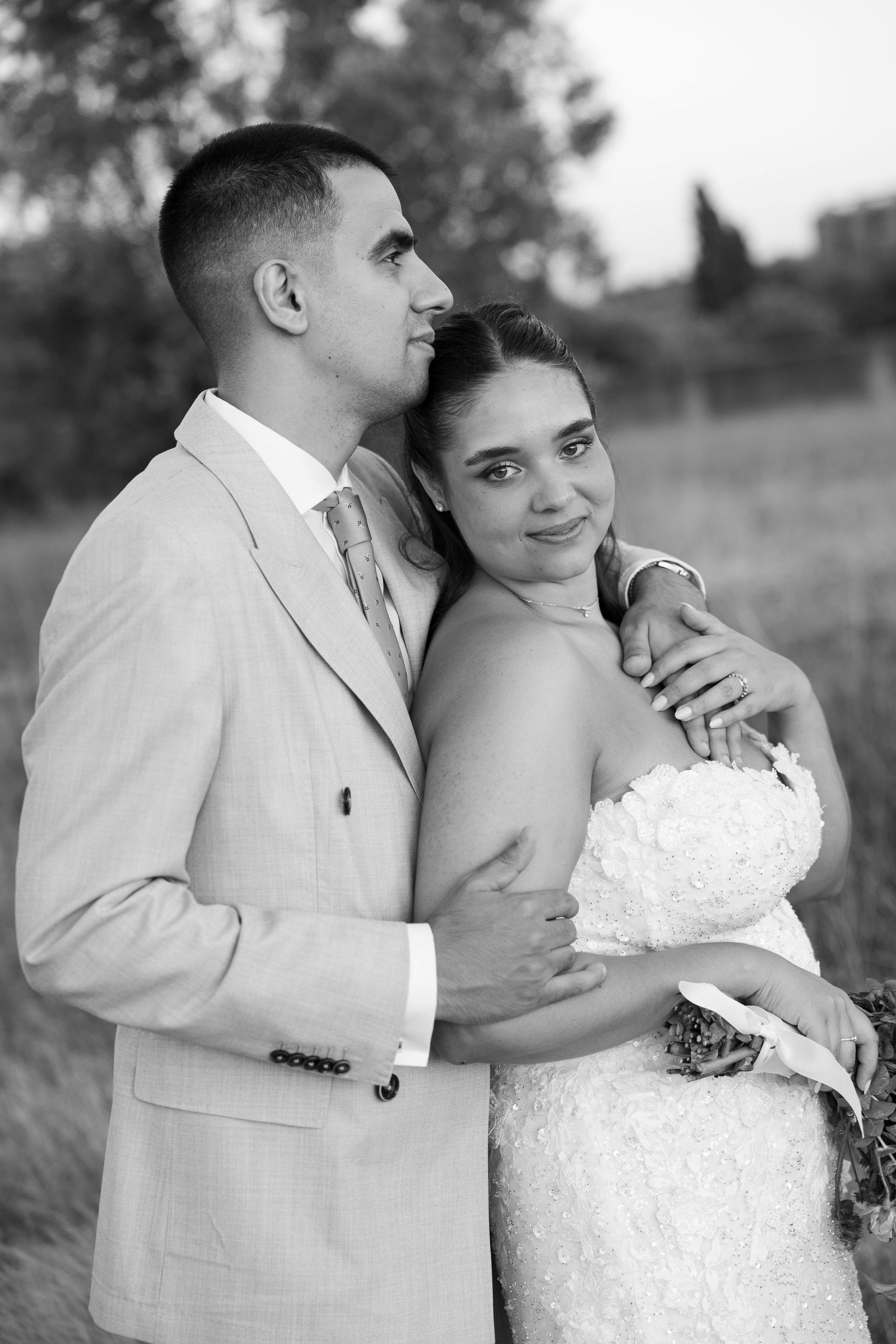 Black and white photo of a bride and groom embracing outdoors, bride holding white bouquet in elegant lace dress.