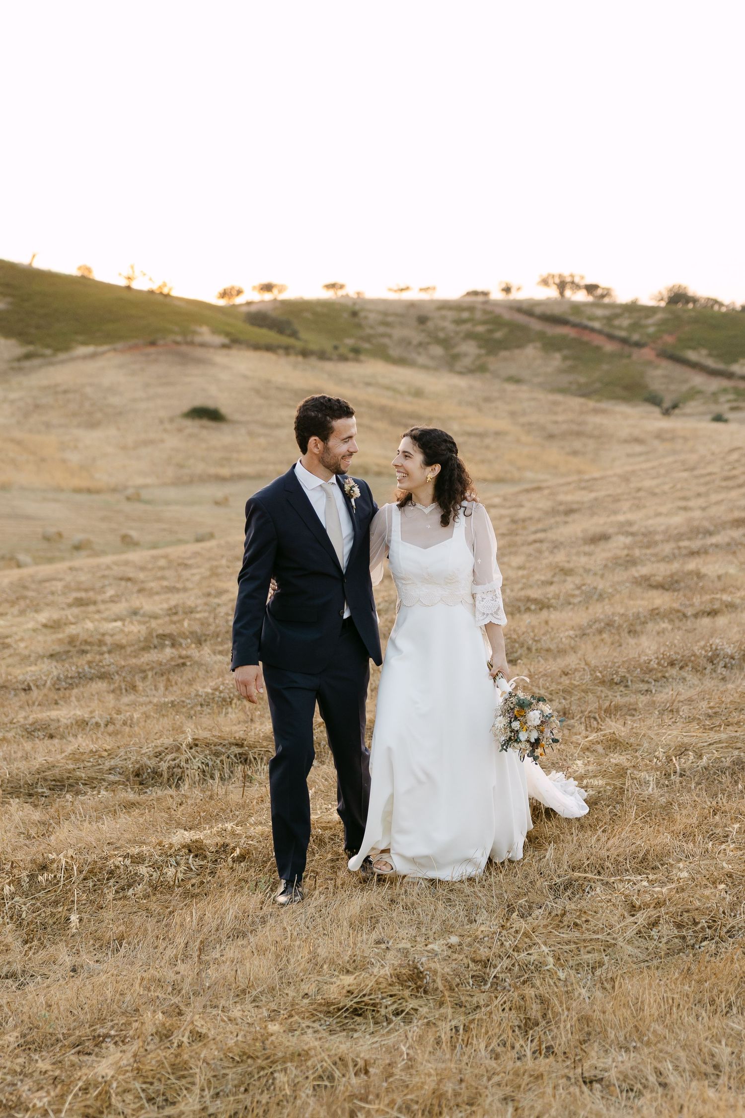 A bride and groom walk through golden rolling hills in Alentejo, smiling at each other, she holds a wildflower bouquet in a white gown.