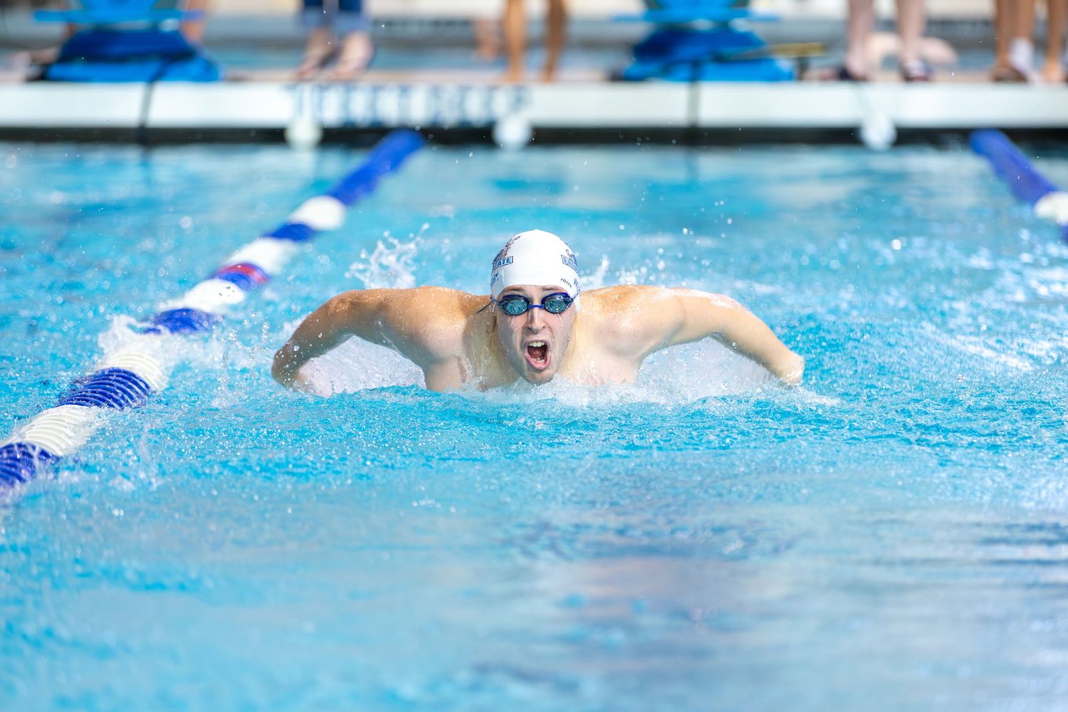 Swimmer performing butterfly stroke in competition pool with blue lane dividers and splashing water.