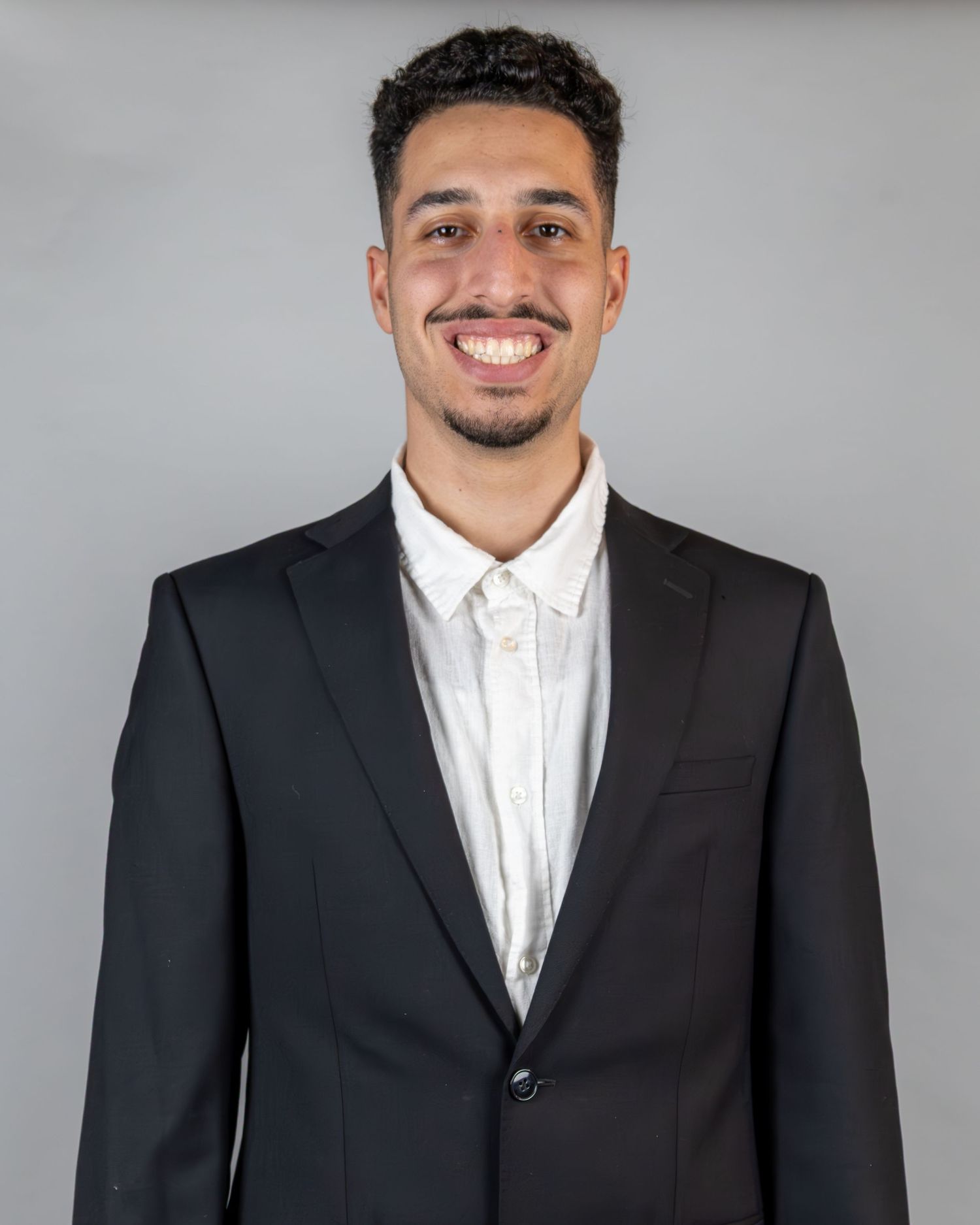 Professional headshot of someone wearing a black suit jacket and white dress shirt against a gray background.