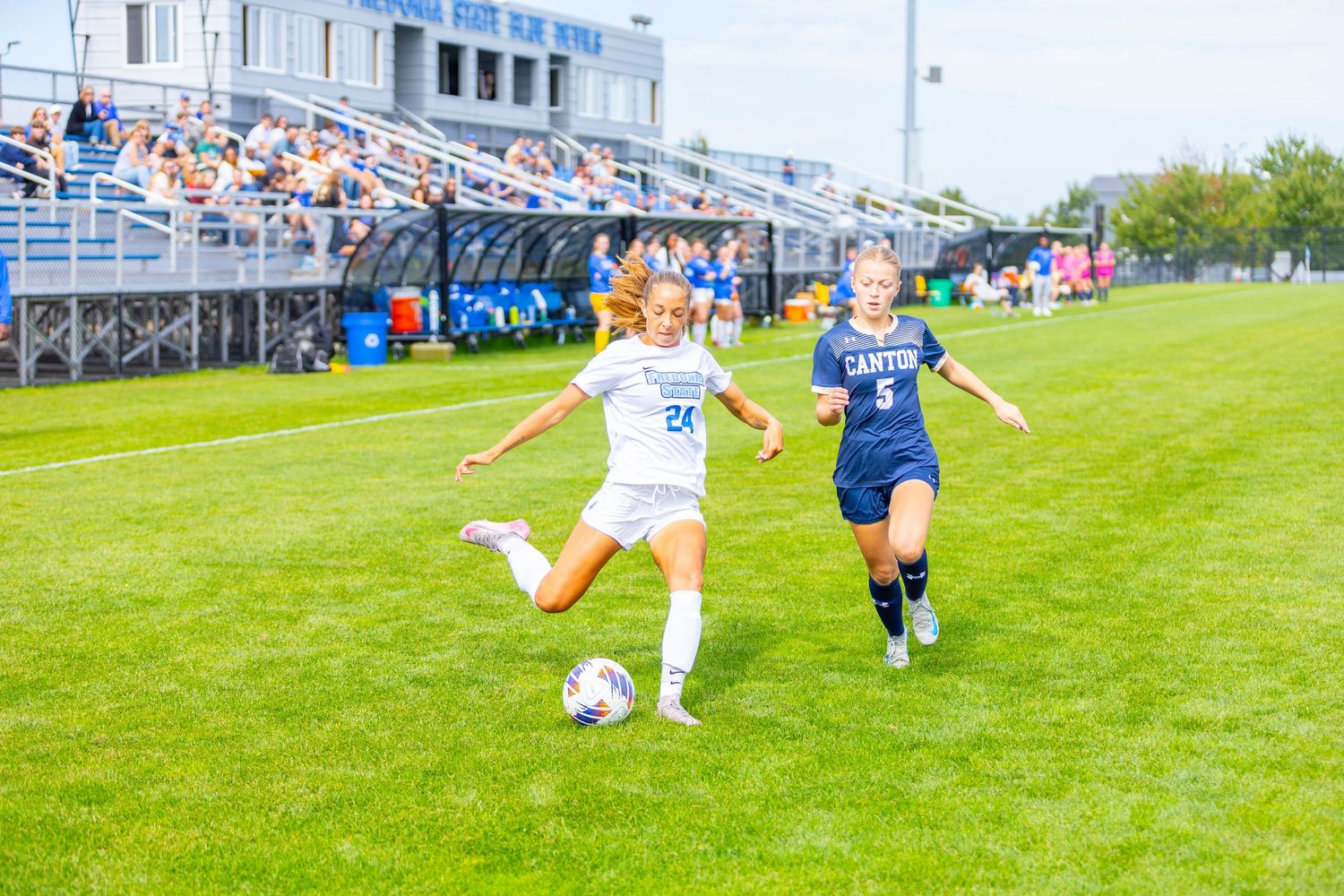 Two soccer players compete for the ball on a grass field with spectator bleachers in the background.