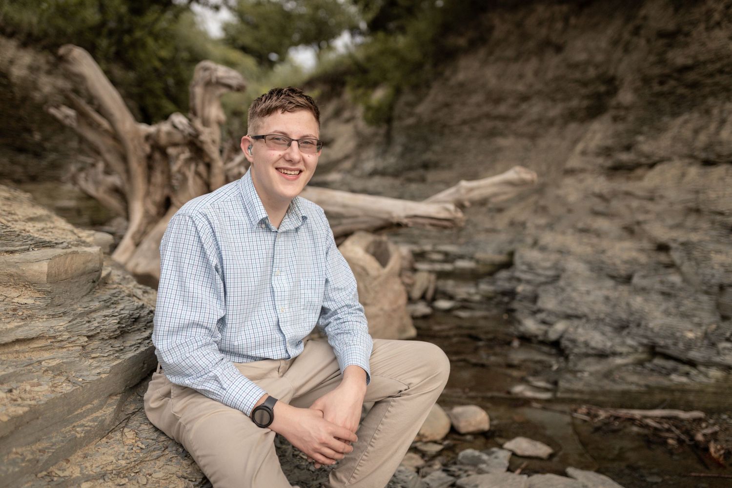A casual portrait of someone sitting on rocky terrain wearing a light blue button-down shirt.
