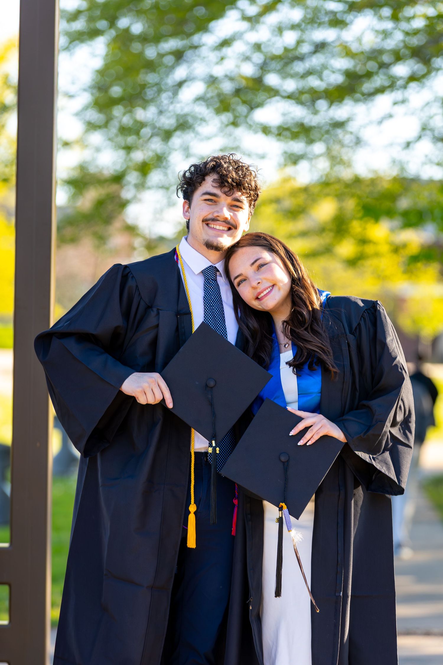 Two smiling graduates in black caps and gowns pose together outdoors on a sunny day.