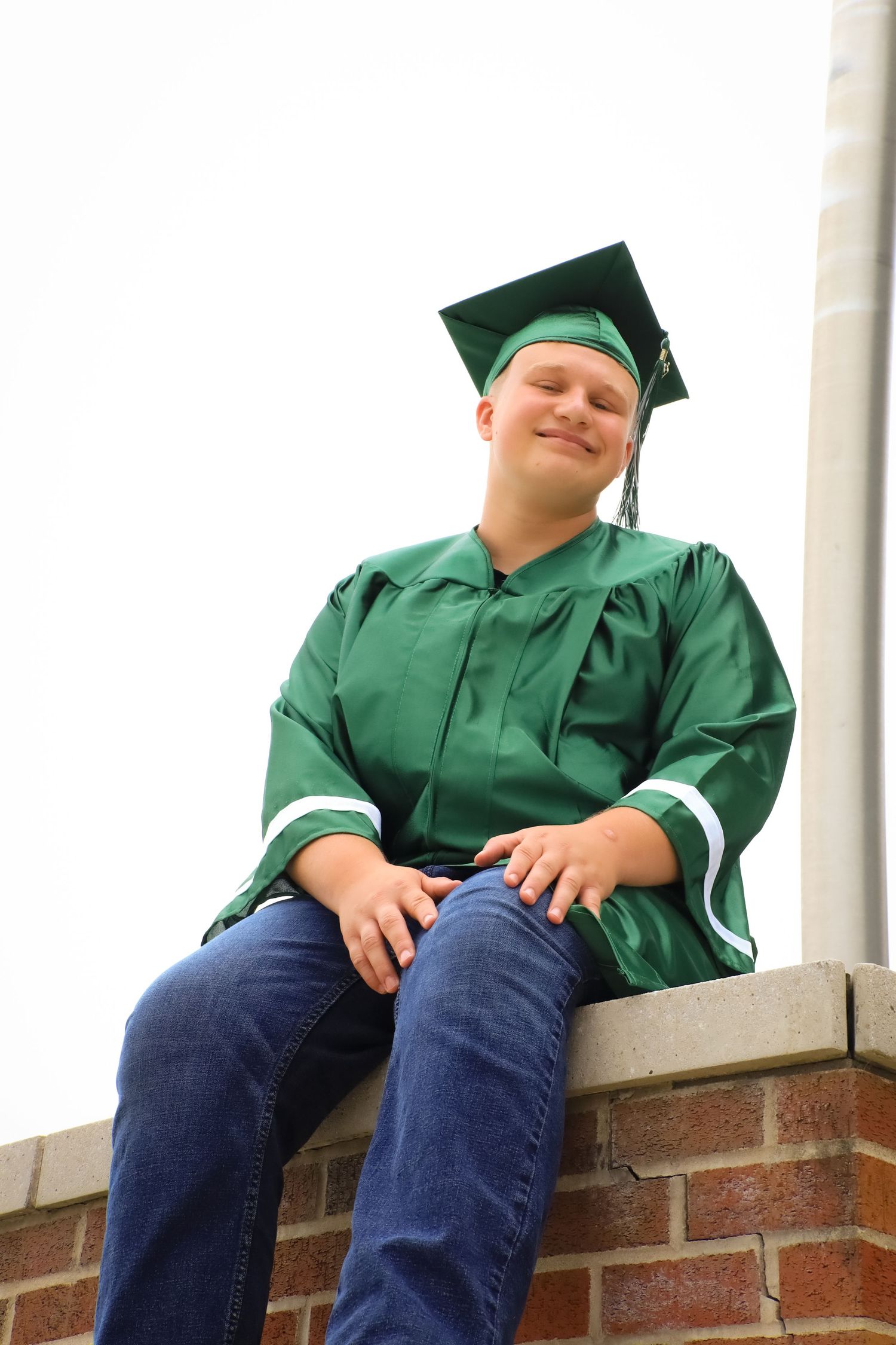 Graduate in green cap and gown sits casually on brick wall during outdoor photo session.
