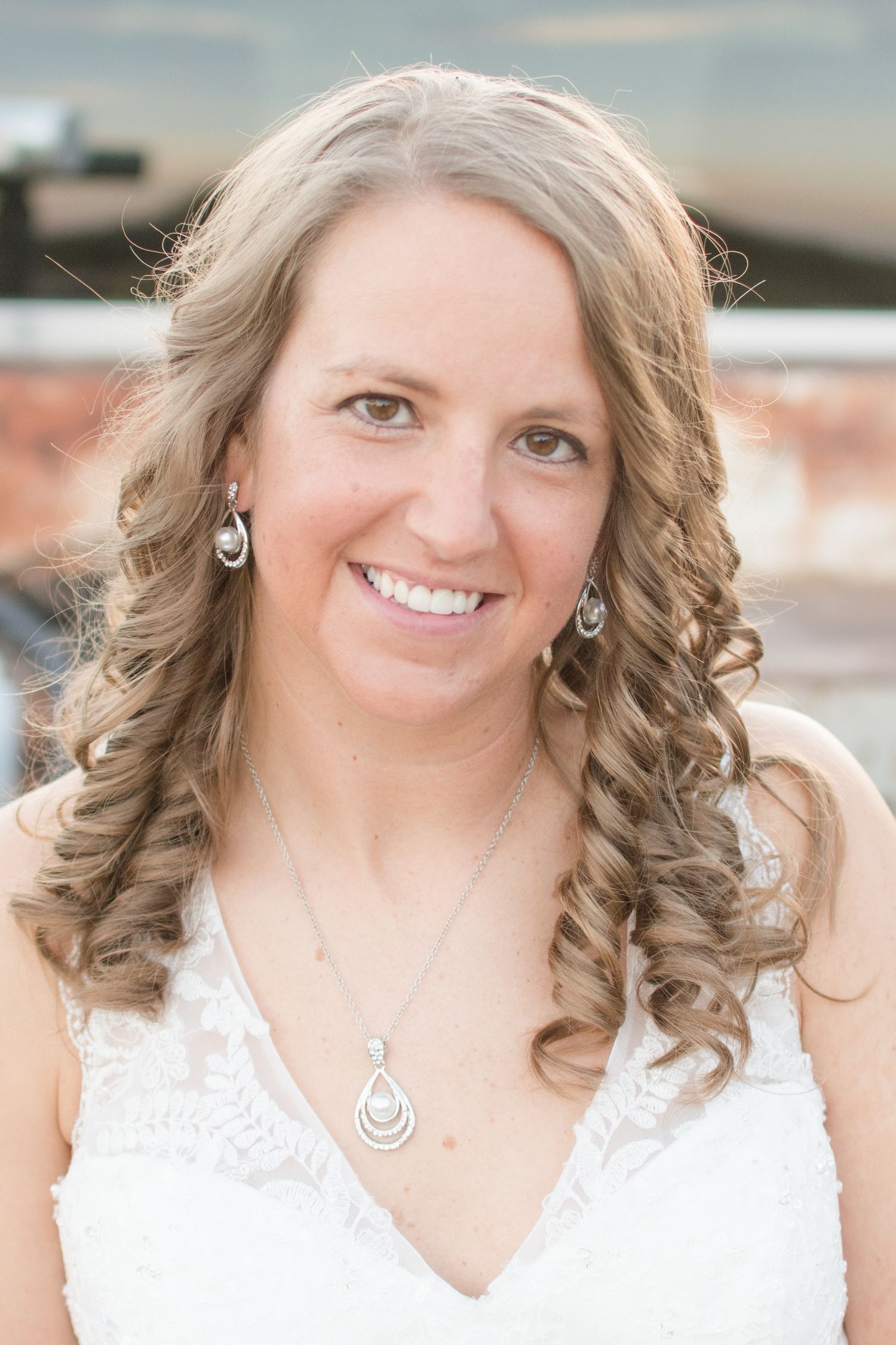 A portrait of someone in a white lace wedding dress with curled hair wearing matching teardrop necklace and earrings.