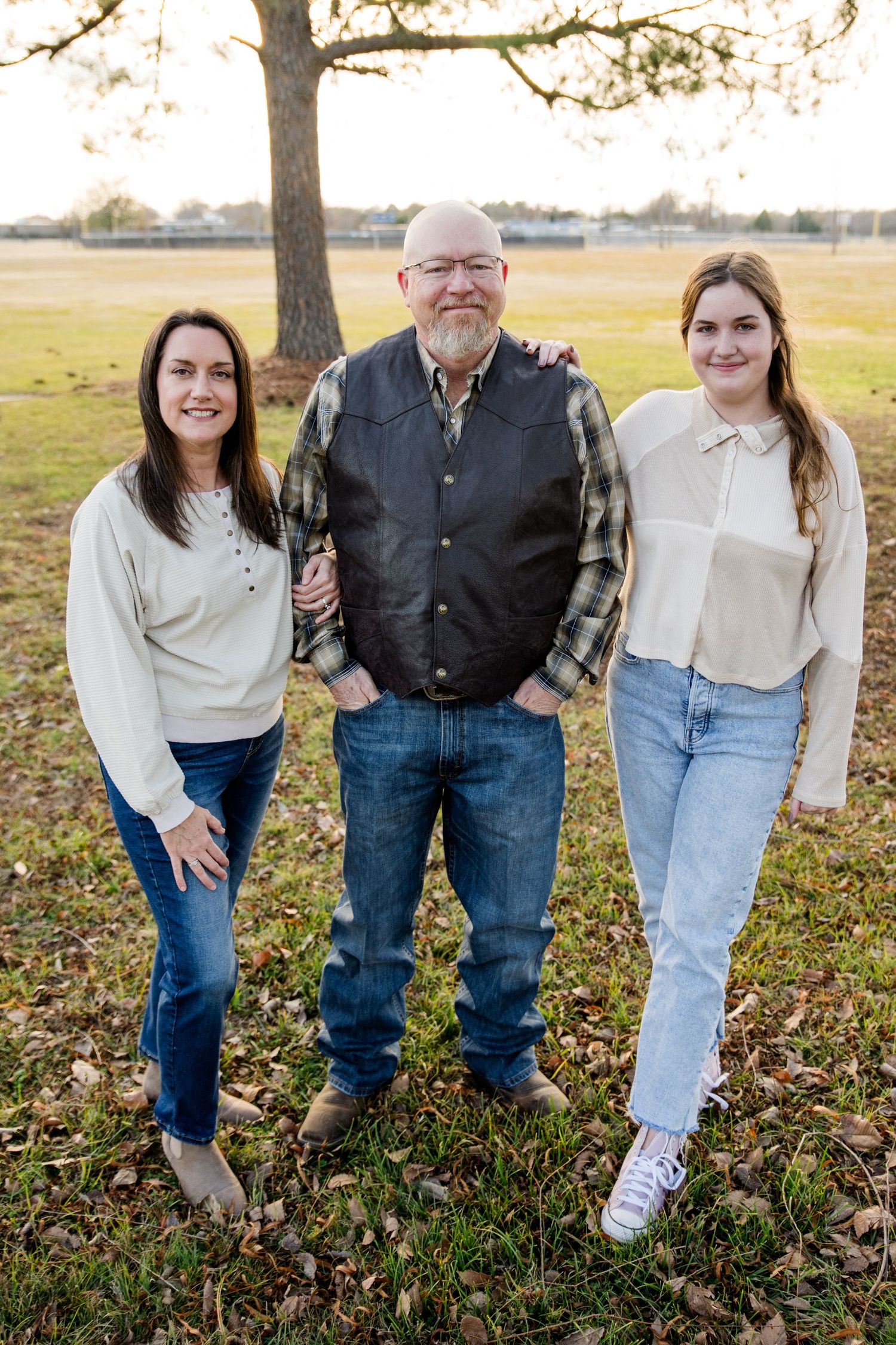 Three people stand together on grass during autumn, wearing casual clothing and smiling outdoors.