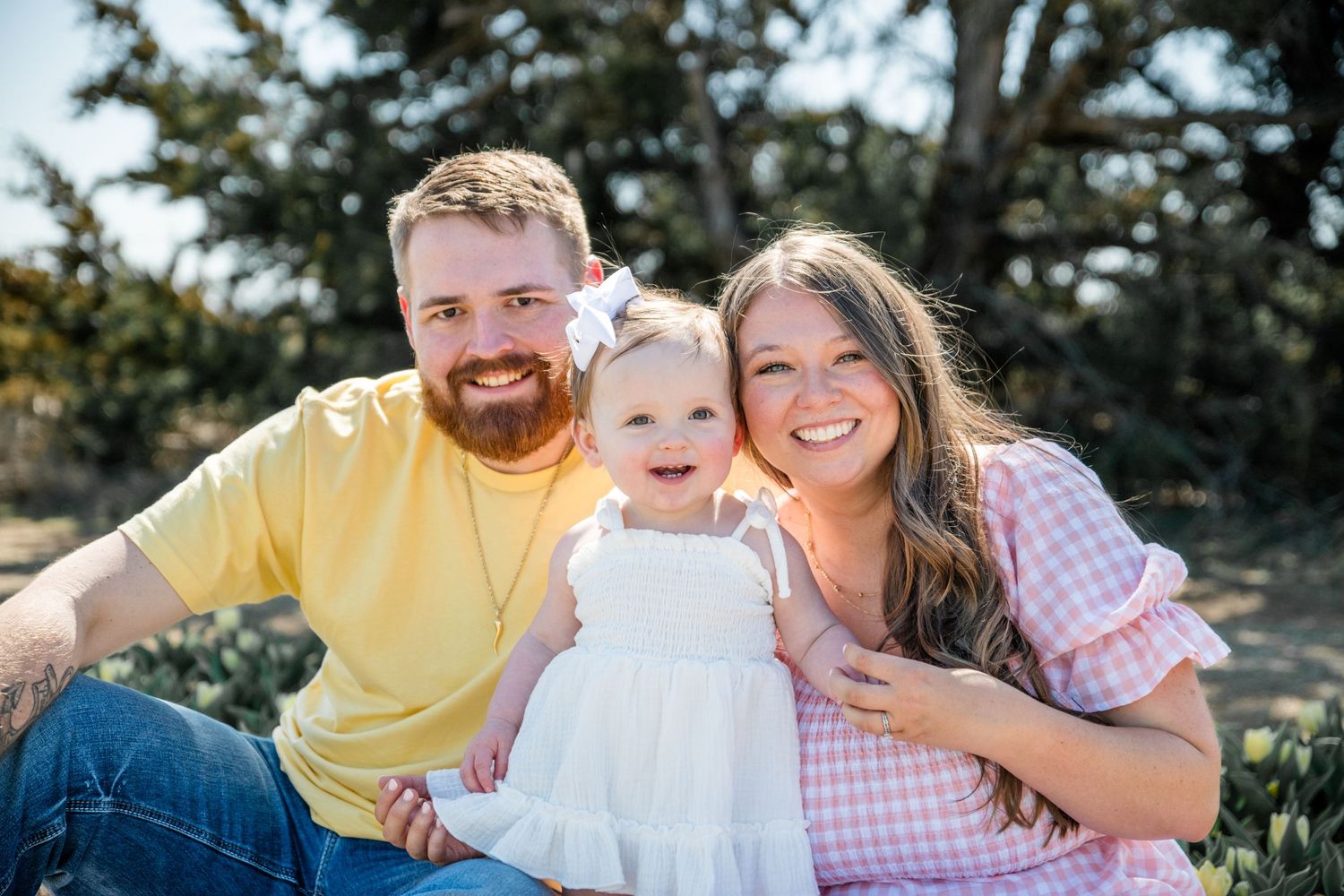 A joyful family in casual attire sits together outdoors on a sunny day amongst greenery.