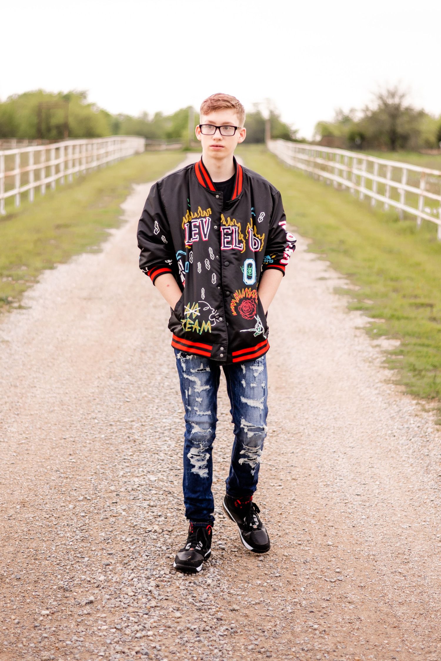 Young person wearing a varsity jacket and ripped jeans stands on a dirt path between white fences.