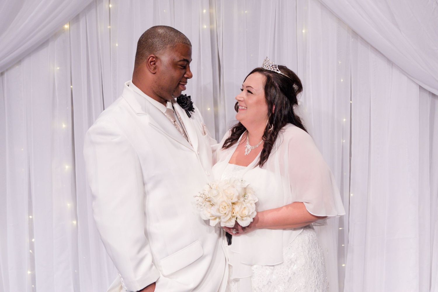A bride in a white dress holds a cream bouquet while standing next to her groom in a white suit at a wedding ceremony.
