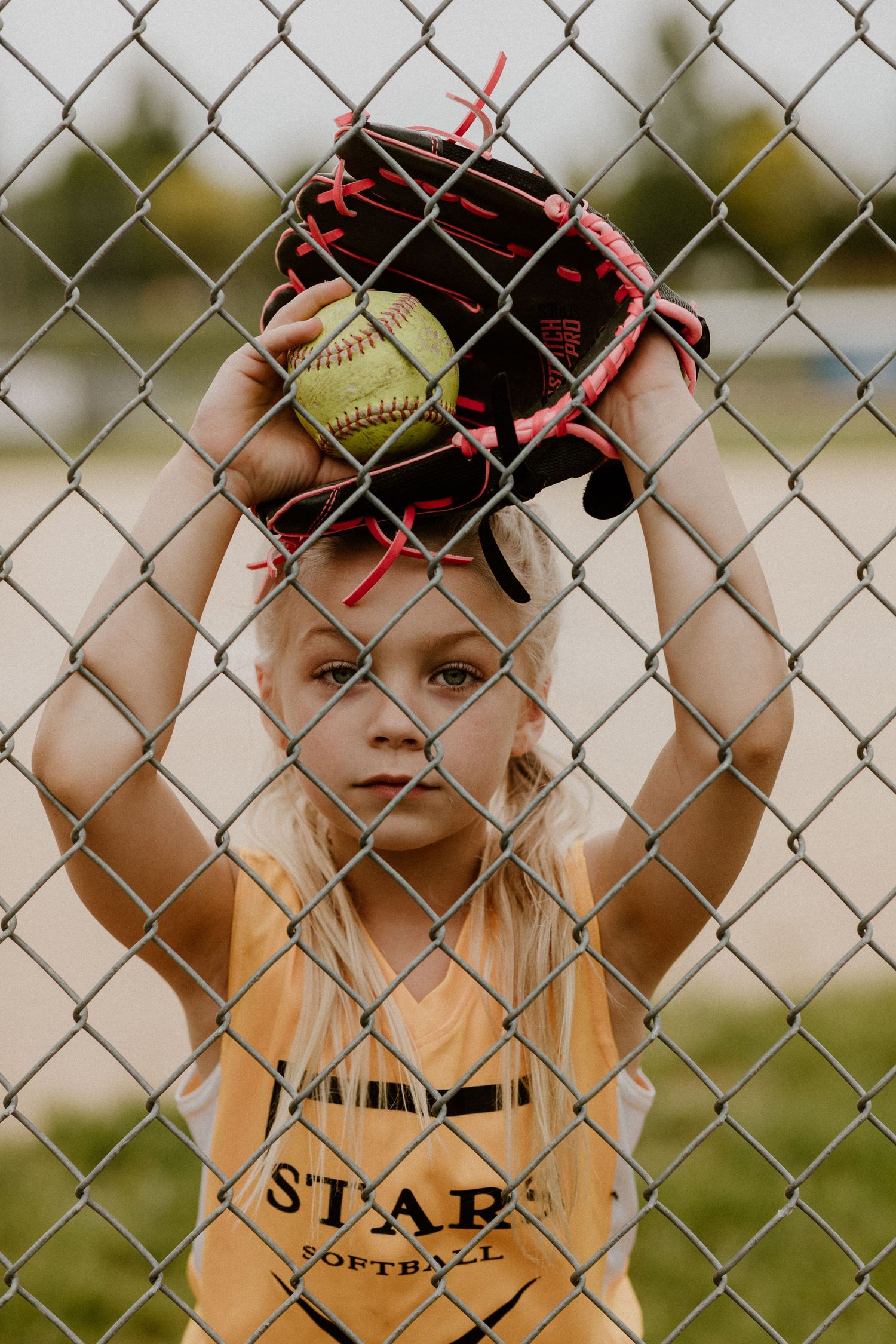 Young softball player in yellow uniform holding glove against chain link fence during practice.