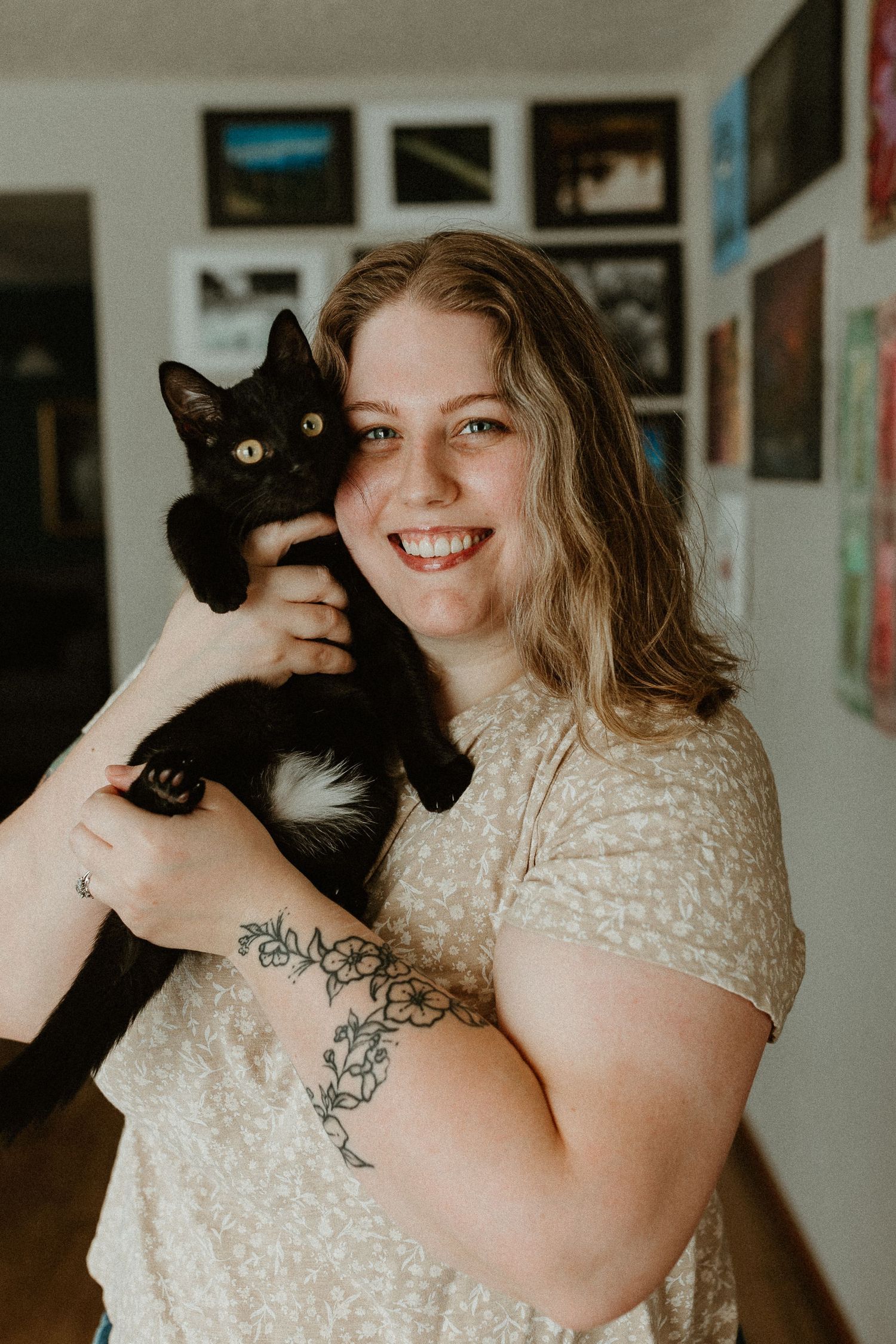 Photographer holding black cat while posing in front of gallery wall with various framed photographs.