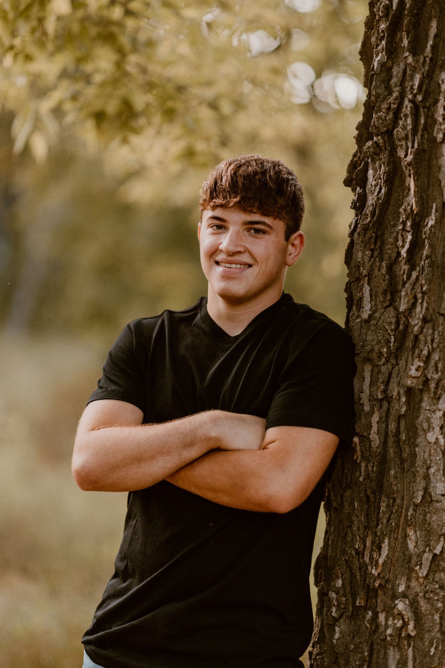 A casual outdoor portrait against a tree trunk with soft bokeh lighting in the background.