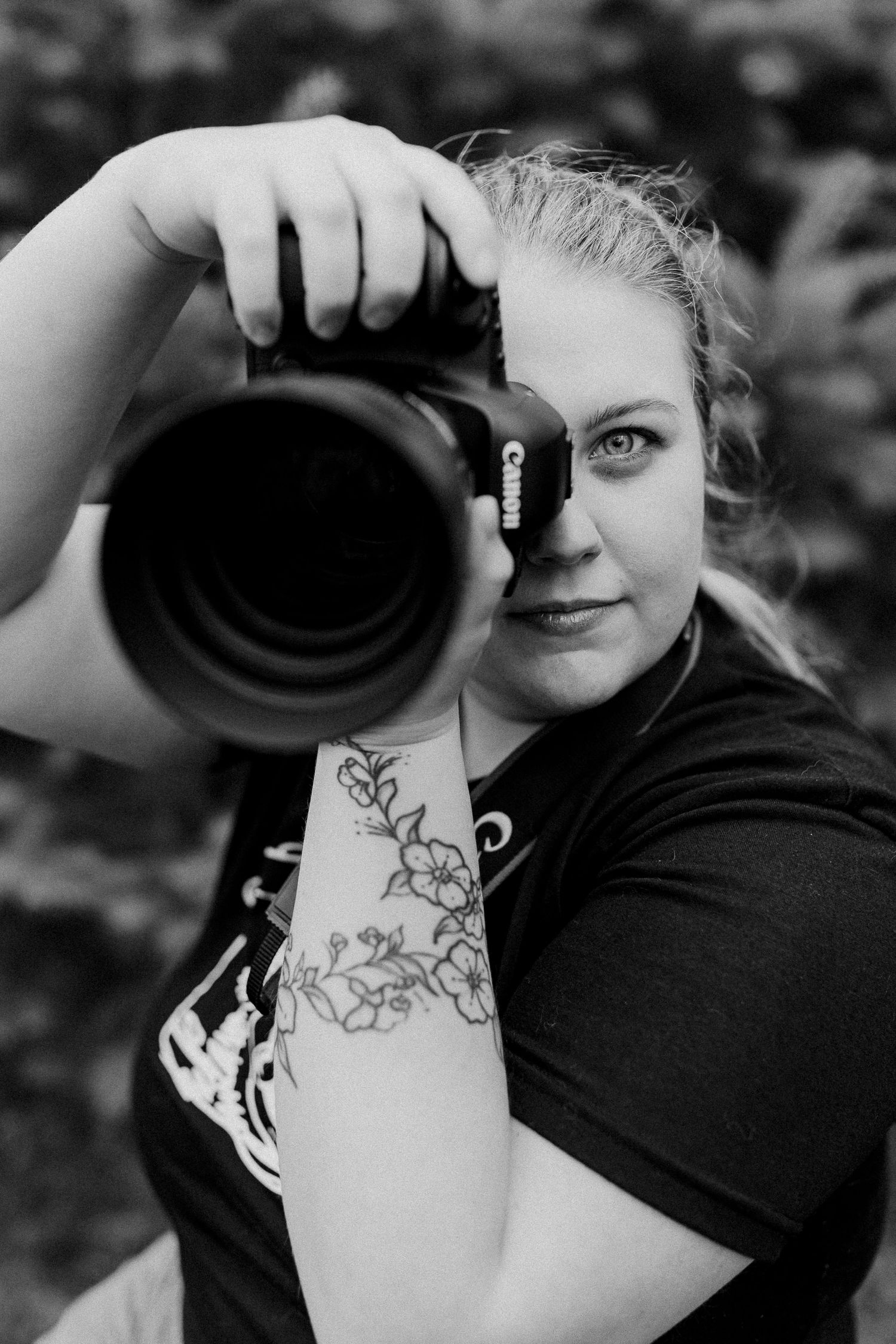 Black and white close-up of photographer with floral arm tattoo holding professional camera.