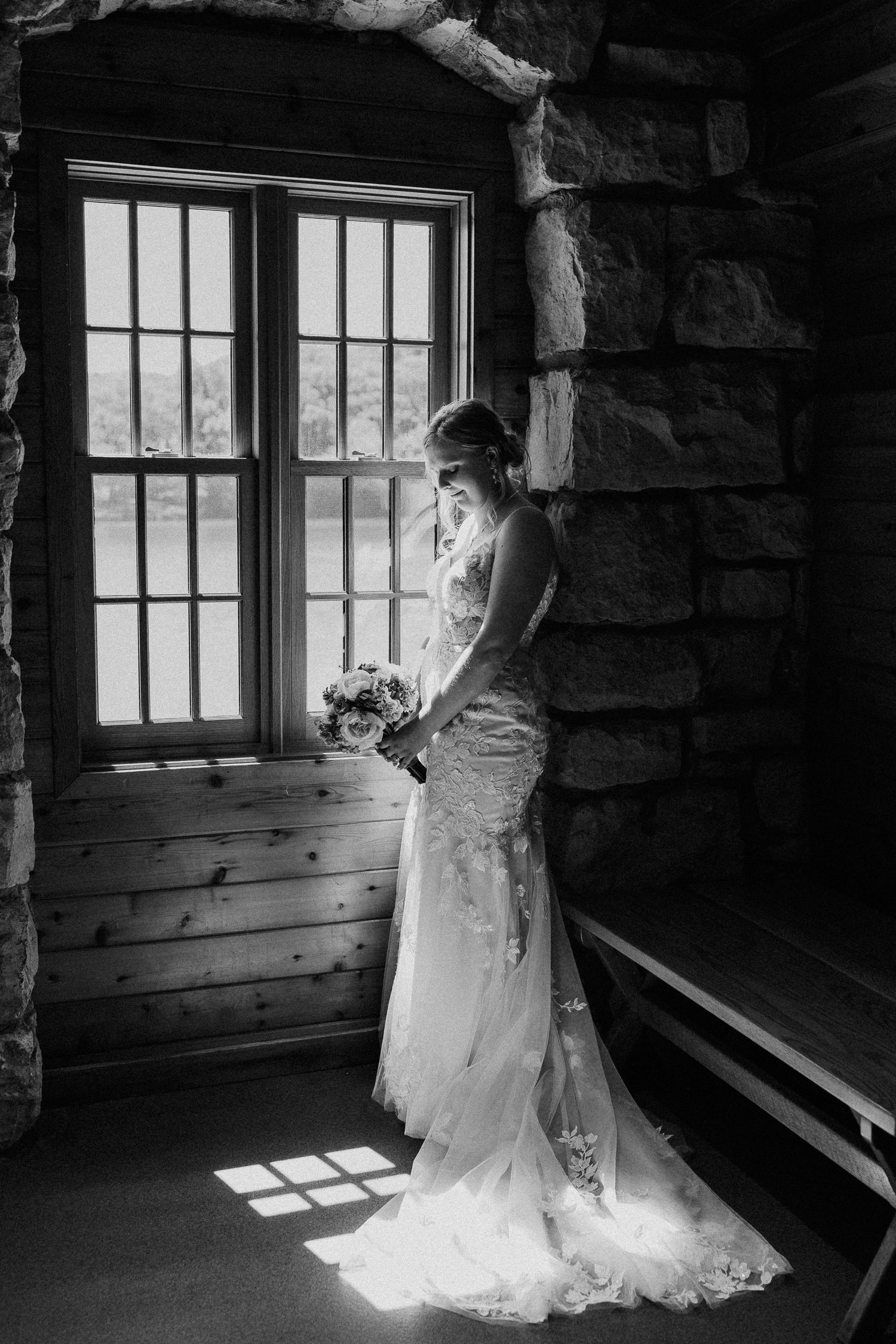 A bride in a lace gown stands by a window in dramatic black and white.