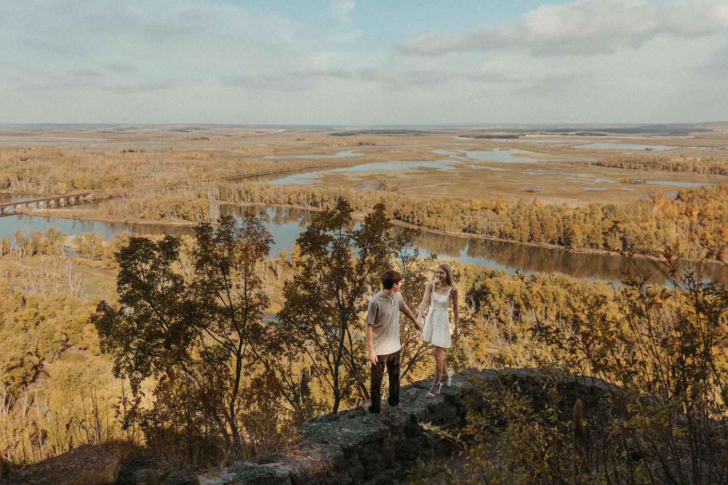 A couple stands on a scenic overlook with fall foliage and water views.