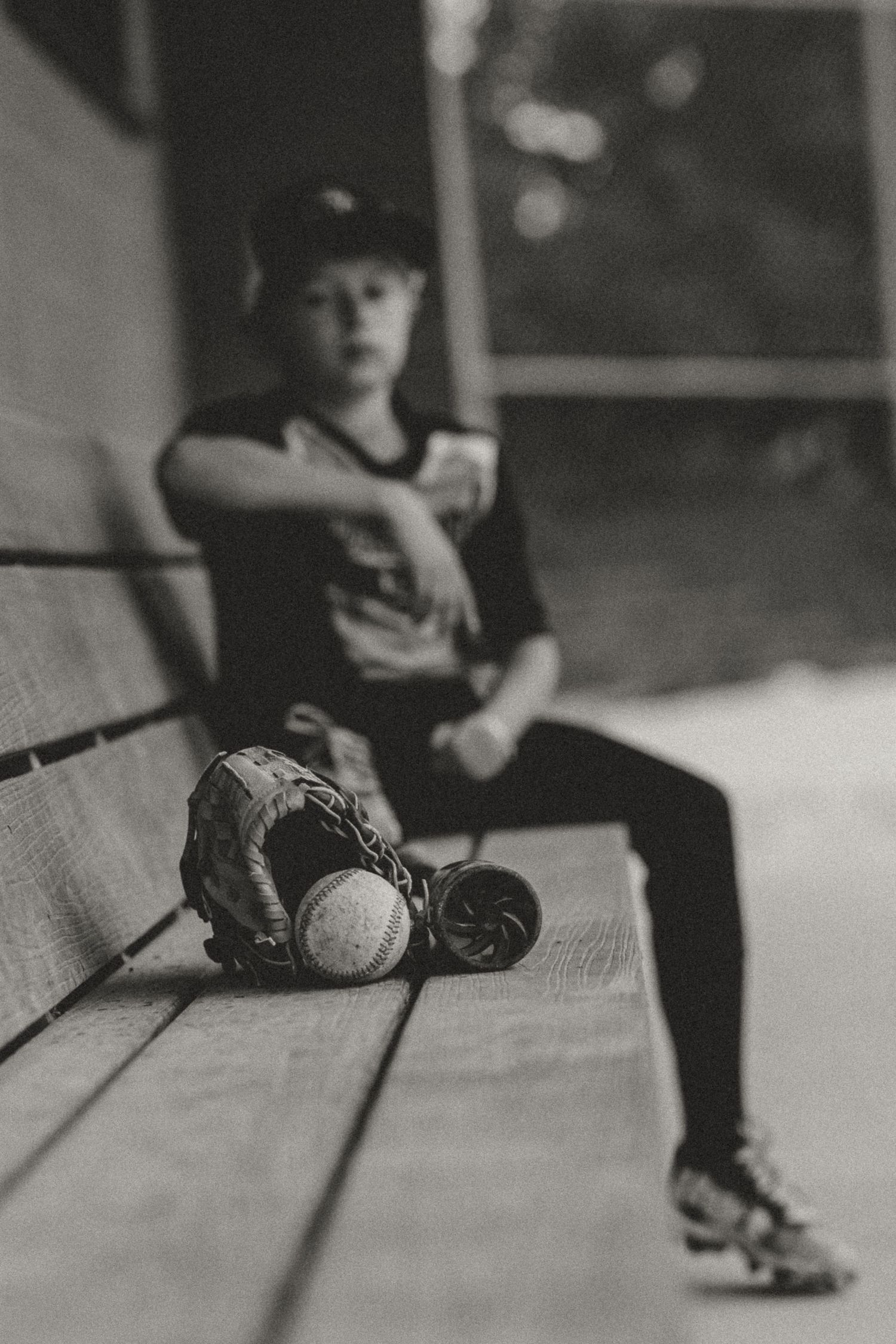 Black and white image of baseball gear on wooden bench with player sitting in background.