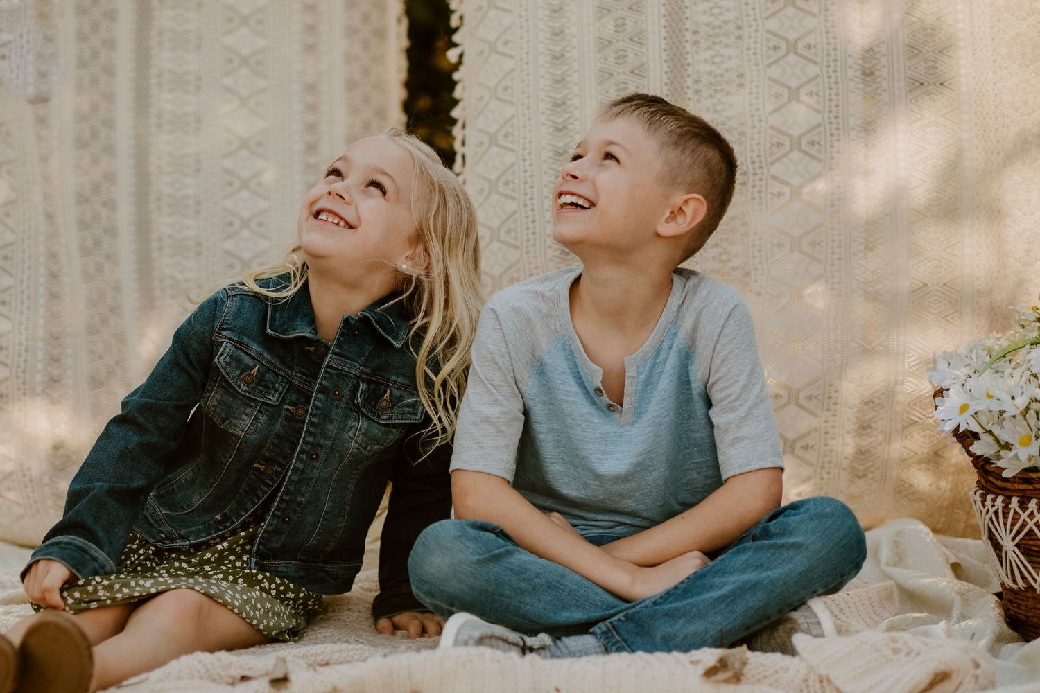 Two children laugh while sitting on a blanket against a white backdrop.