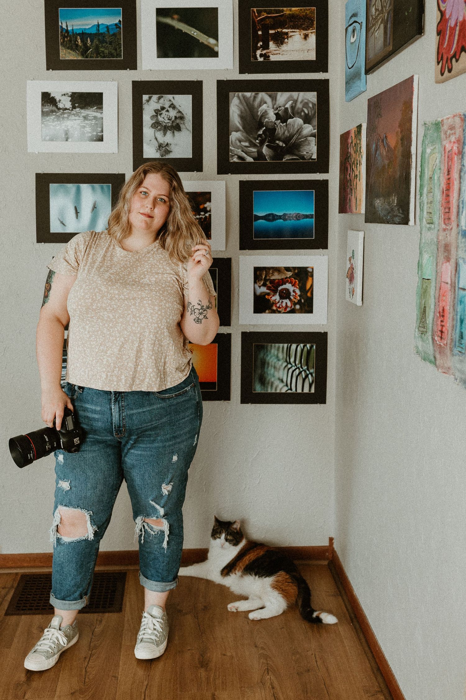 Photographer with camera standing by photo wall while black and white cat rests in corner.