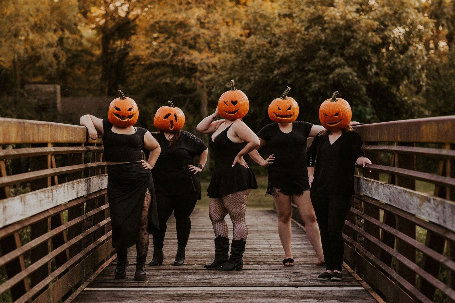 Group wearing black dresses and pumpkin heads pose playfully on wooden bridge.