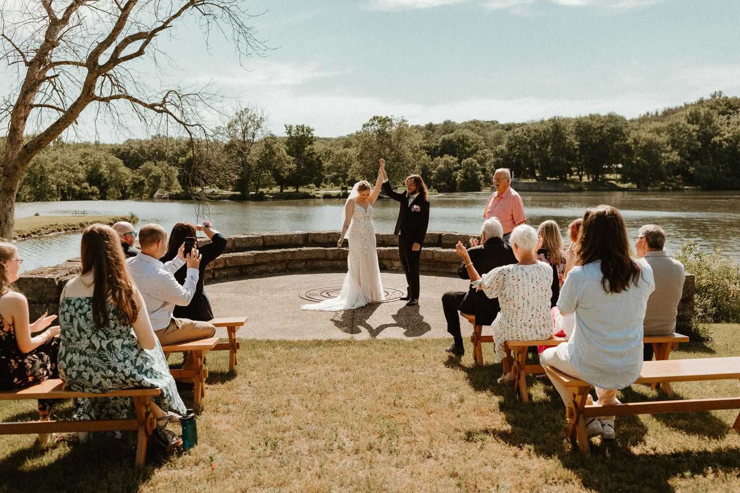 An outdoor wedding ceremony with guests seated on wooden benches by a lake.