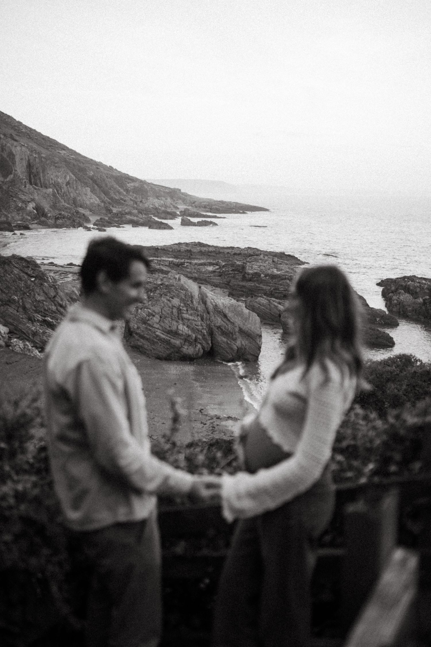 A romantic black and white coastal scene with a couple standing together on rocky cliffs overlooking the ocean.