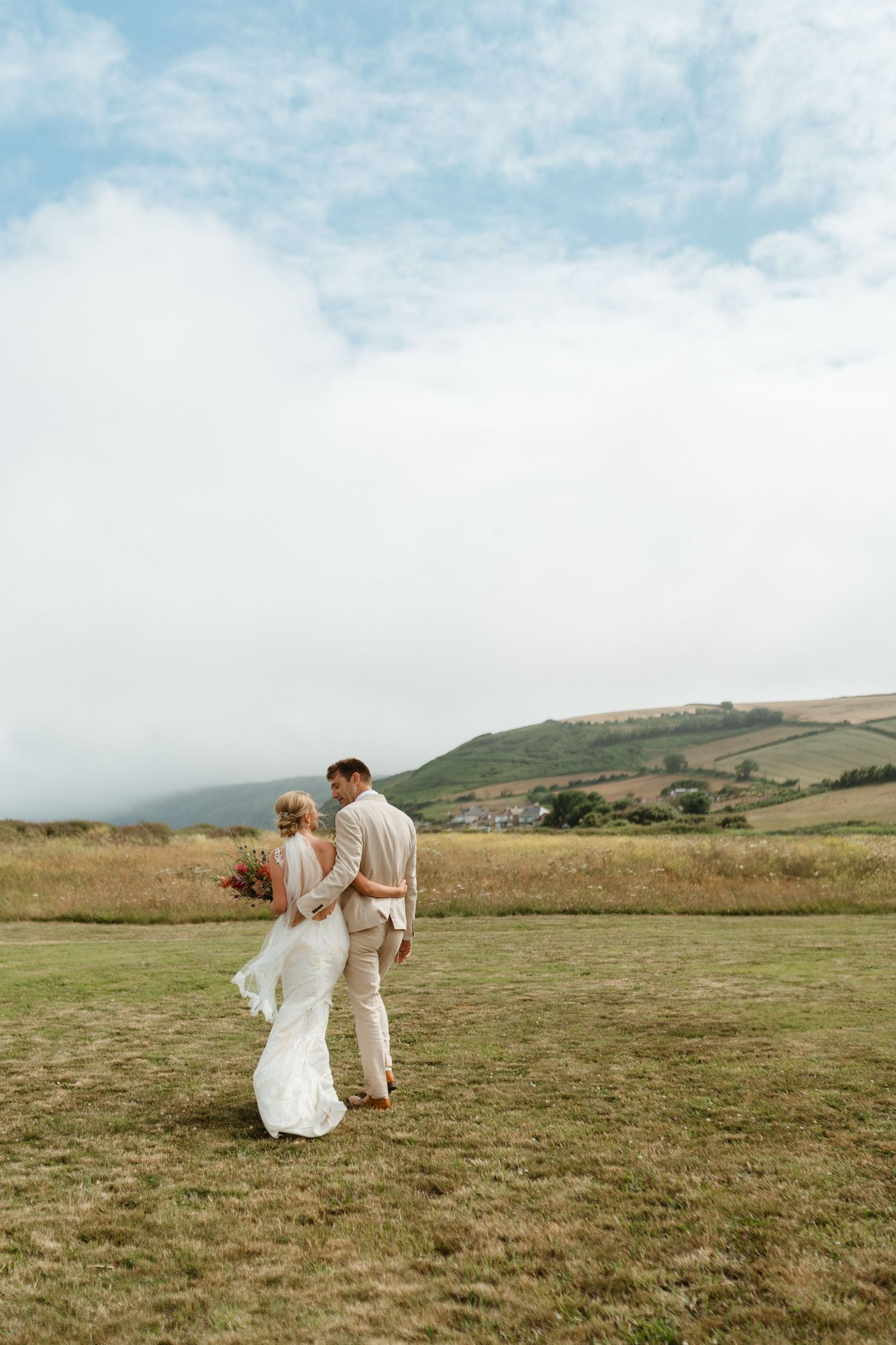 A couple in wedding attire stands embracing in a grassy field with rolling hills and cloudy sky in the background.