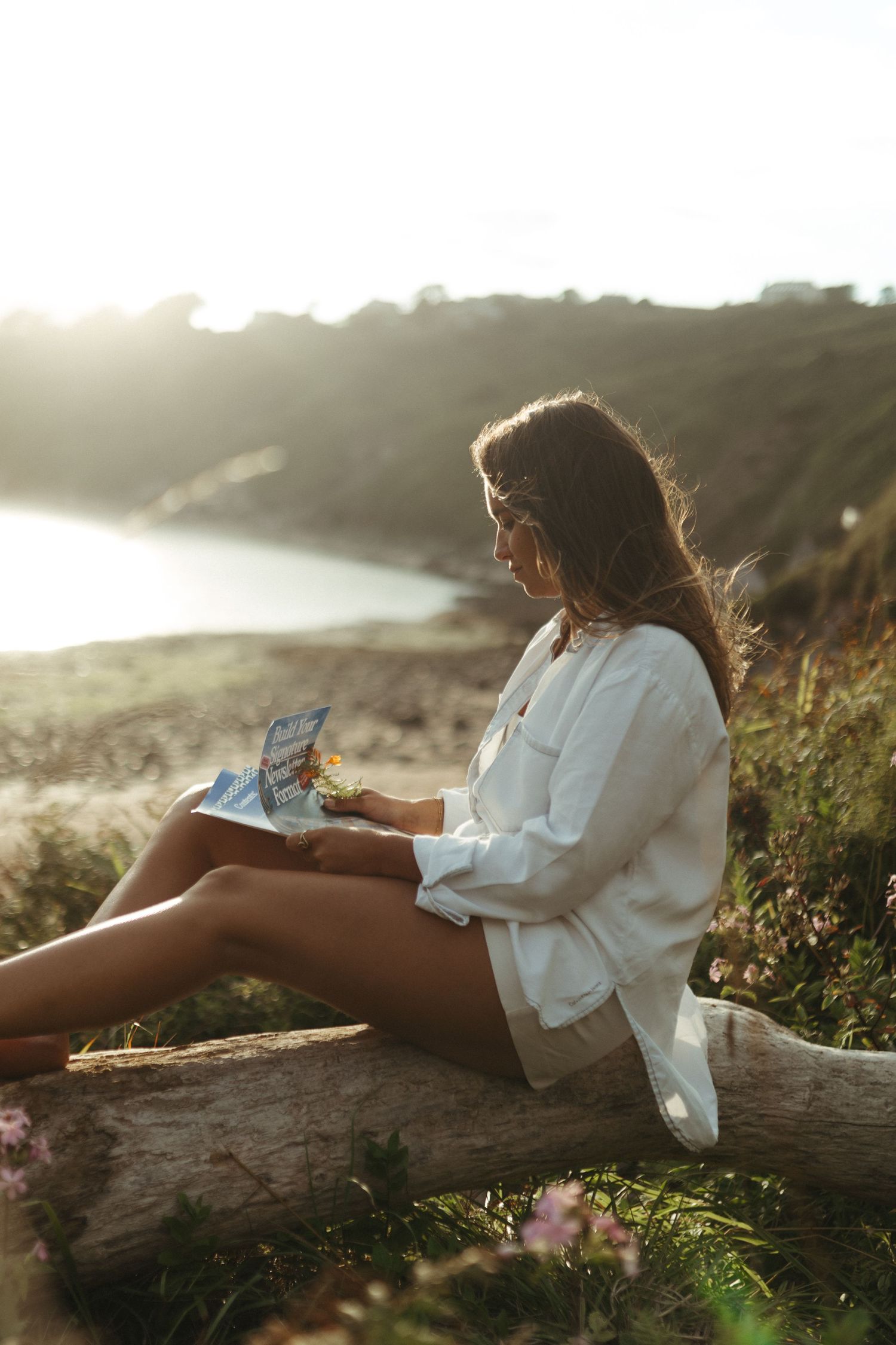 Someone in a white shirt sits on a log at sunset reading a book along a scenic coastal overlook.