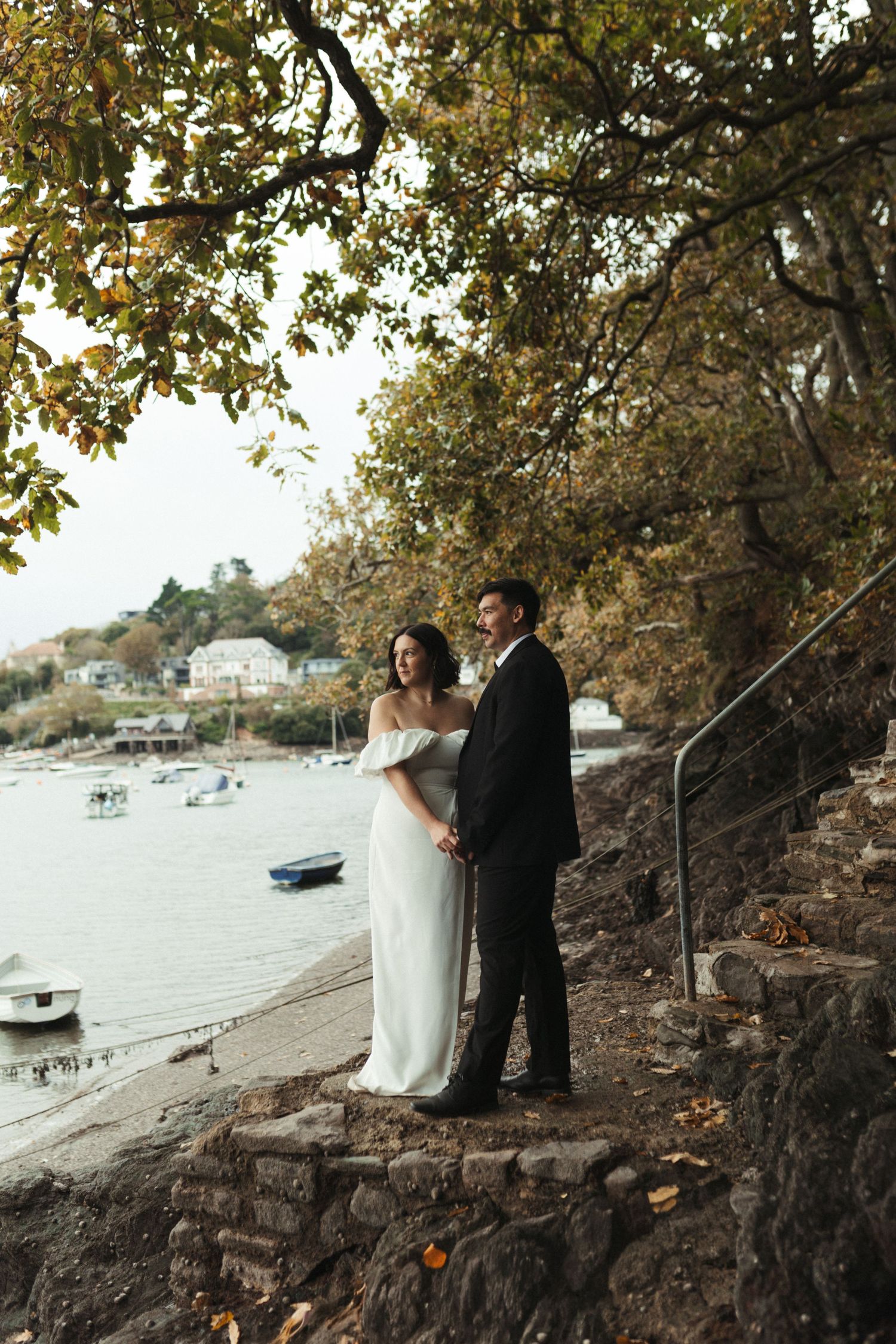 A romantic wedding couple stands by a rocky waterfront with boats in the distance during autumn.