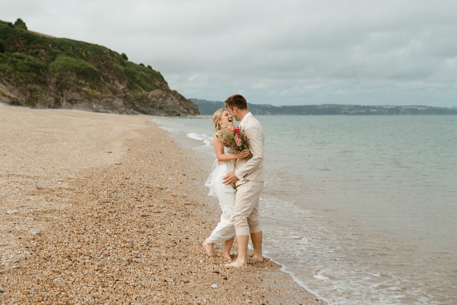 Couple in white clothing embraces intimately on a serene beach with soft waves and rocky cliffs in the background.