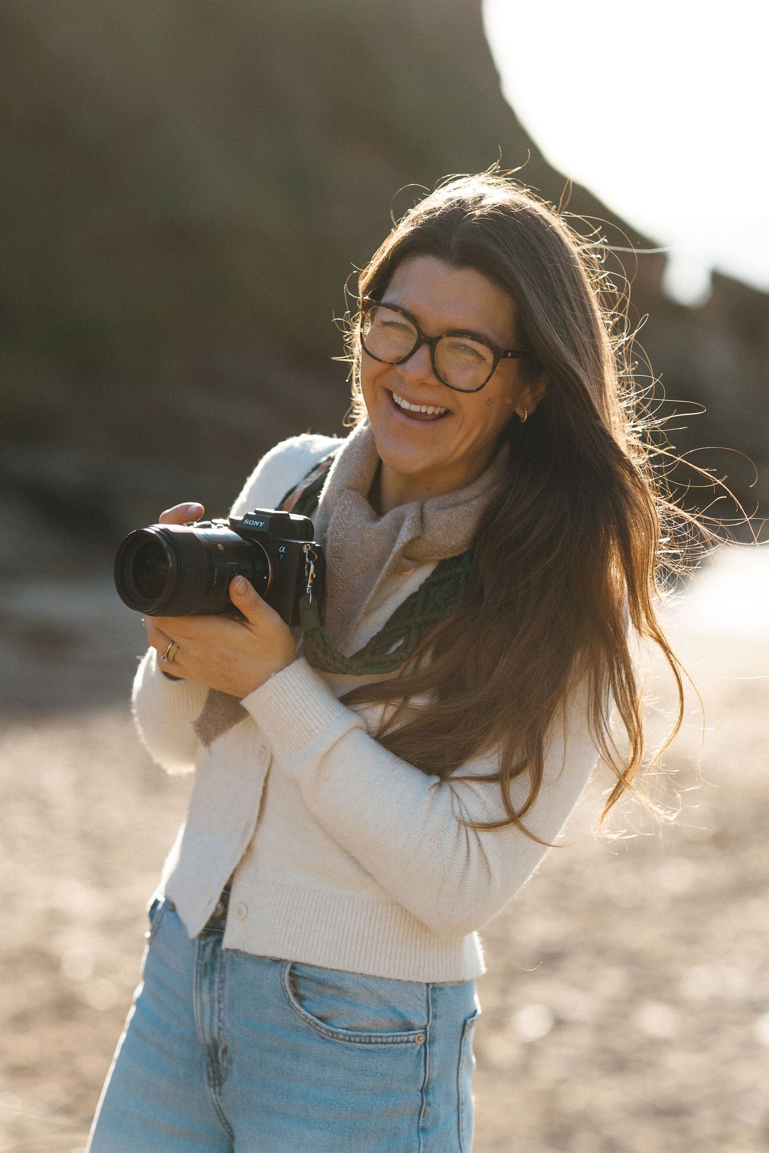 A photographer in glasses and a white sweater laughs while holding a camera during a golden hour photoshoot.