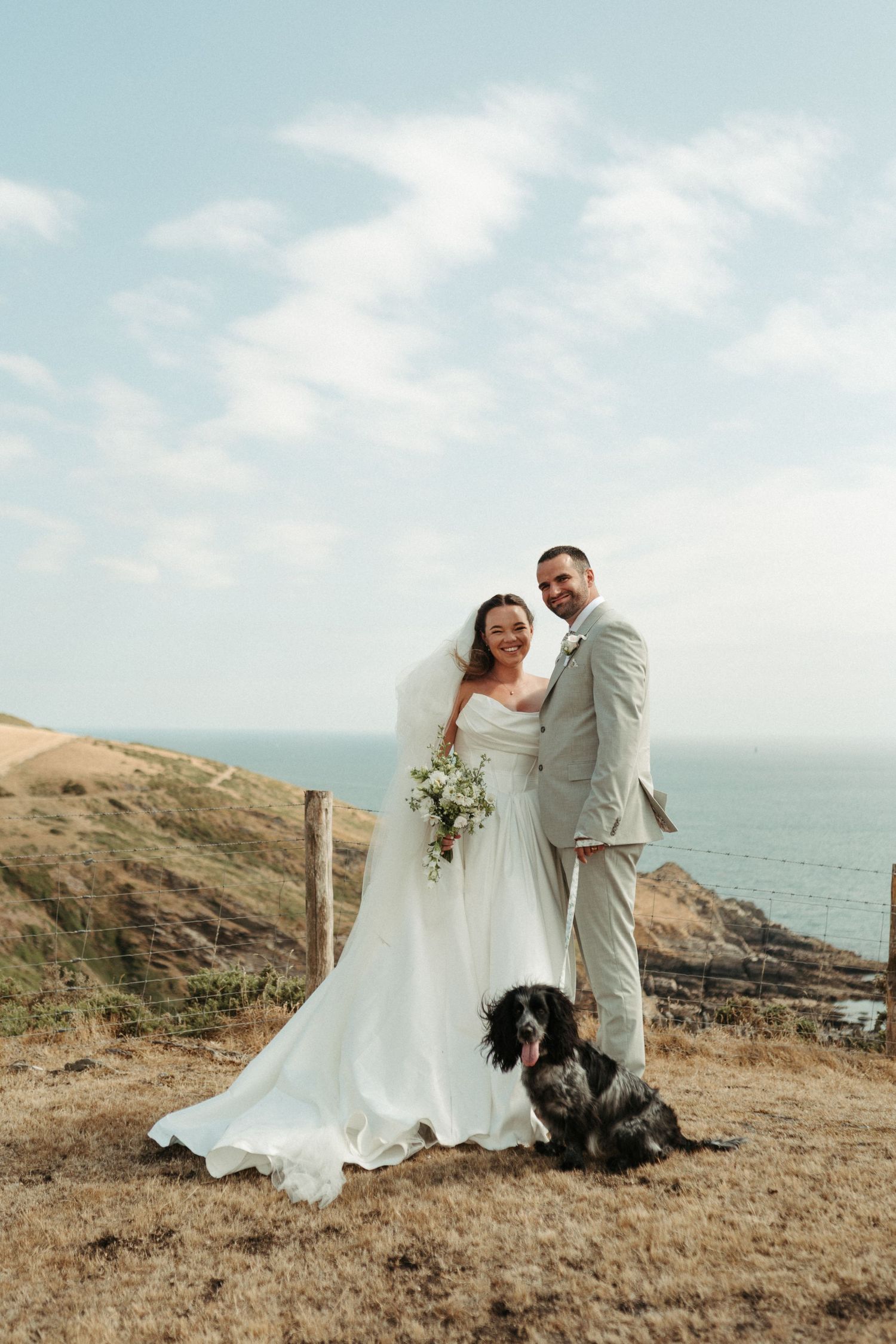 Bride and groom pose with their spaniel dog on a coastal clifftop, bride holding white bouquet in flowing gown.