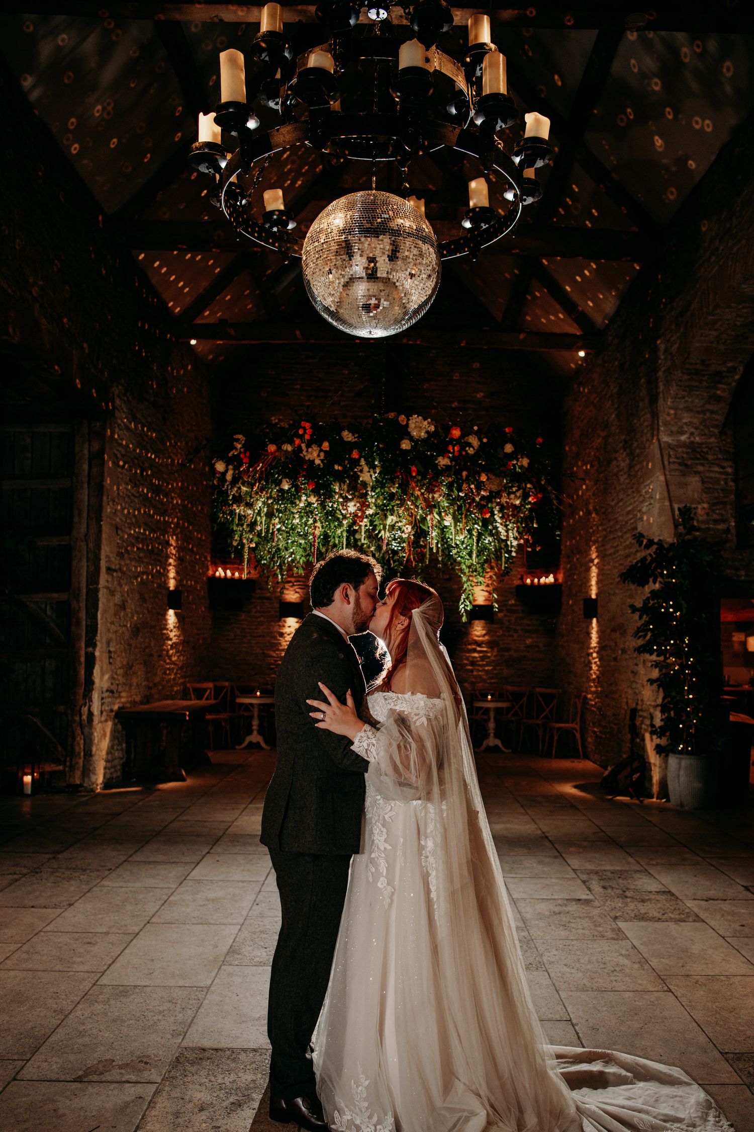 Bride and groom share a romantic moment in a rustic stone barn venue with chandelier and floral installation.