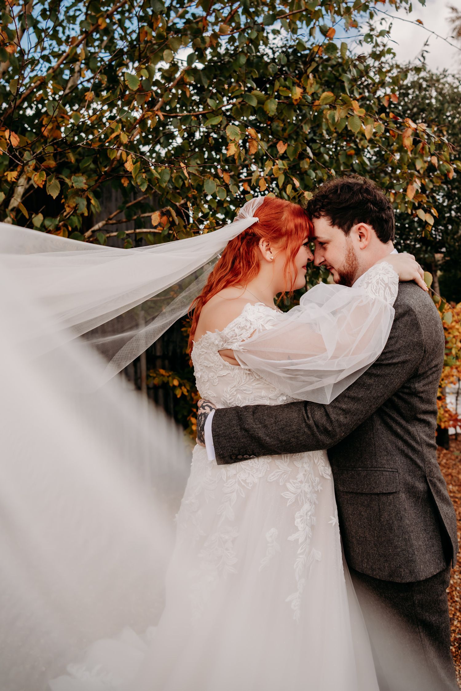 Bride with red hair and flowing veil embraces groom in gray suit outdoors amid autumn foliage.