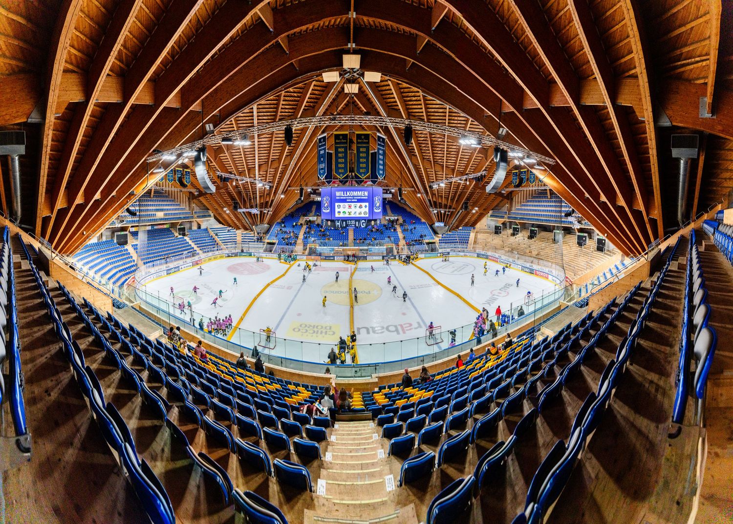 Panoramic view of an empty indoor ice hockey arena with blue seats and curved wooden ceiling beams.