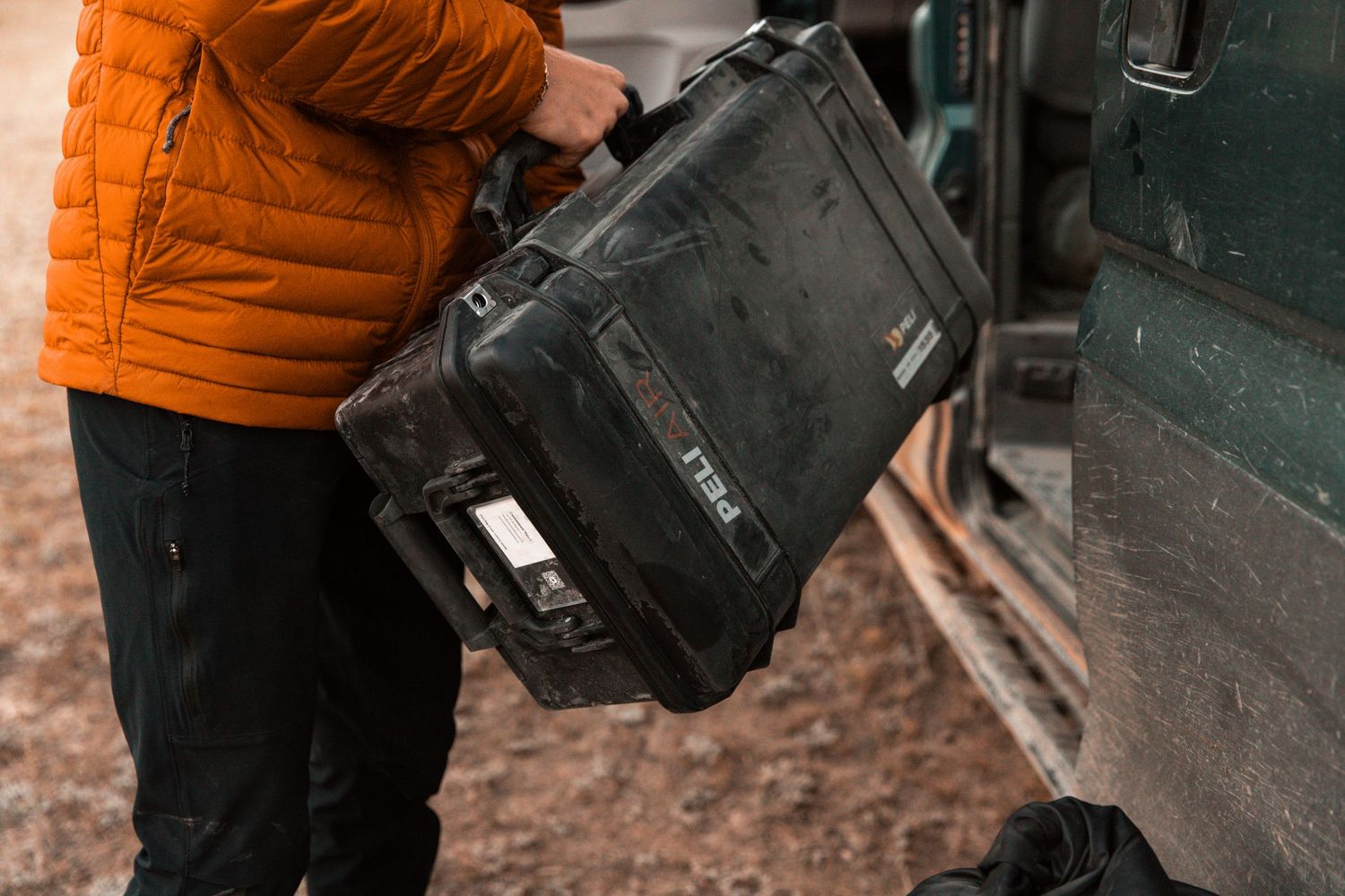 Person in orange puffer jacket carrying a black protective hardshell case next to a vehicle.