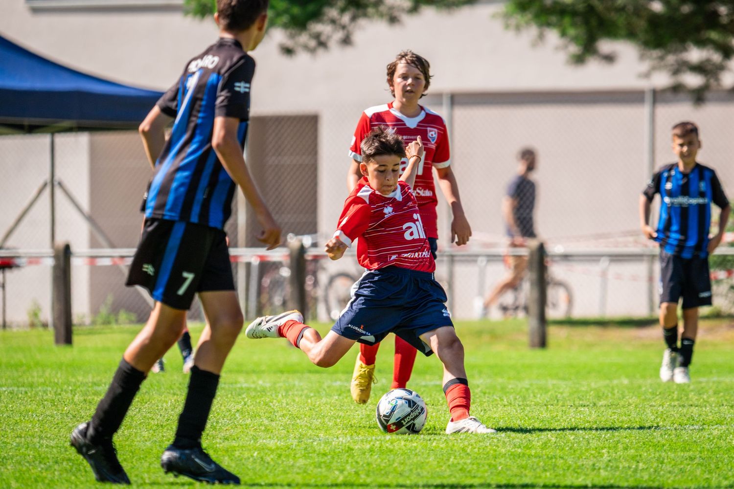 Youth soccer players in red and blue jerseys compete for the ball during an outdoor match on grass field.