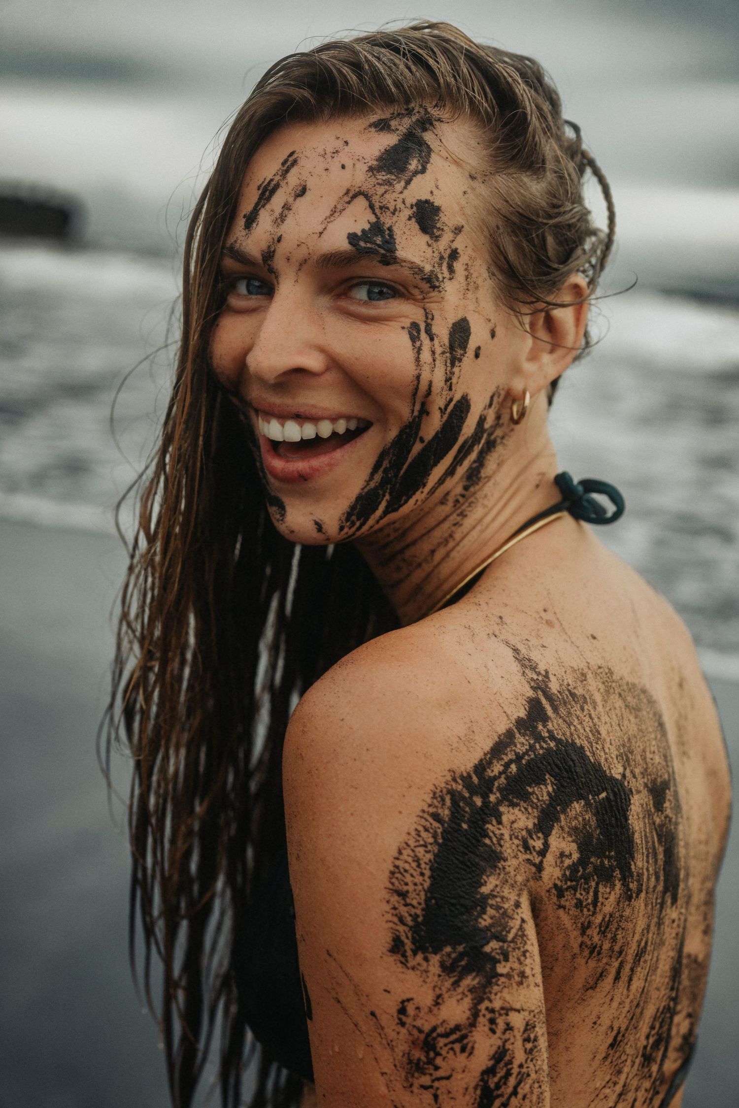 Smiling woman with wet hair and black mud covering her face, neck, and back standing near the ocean.