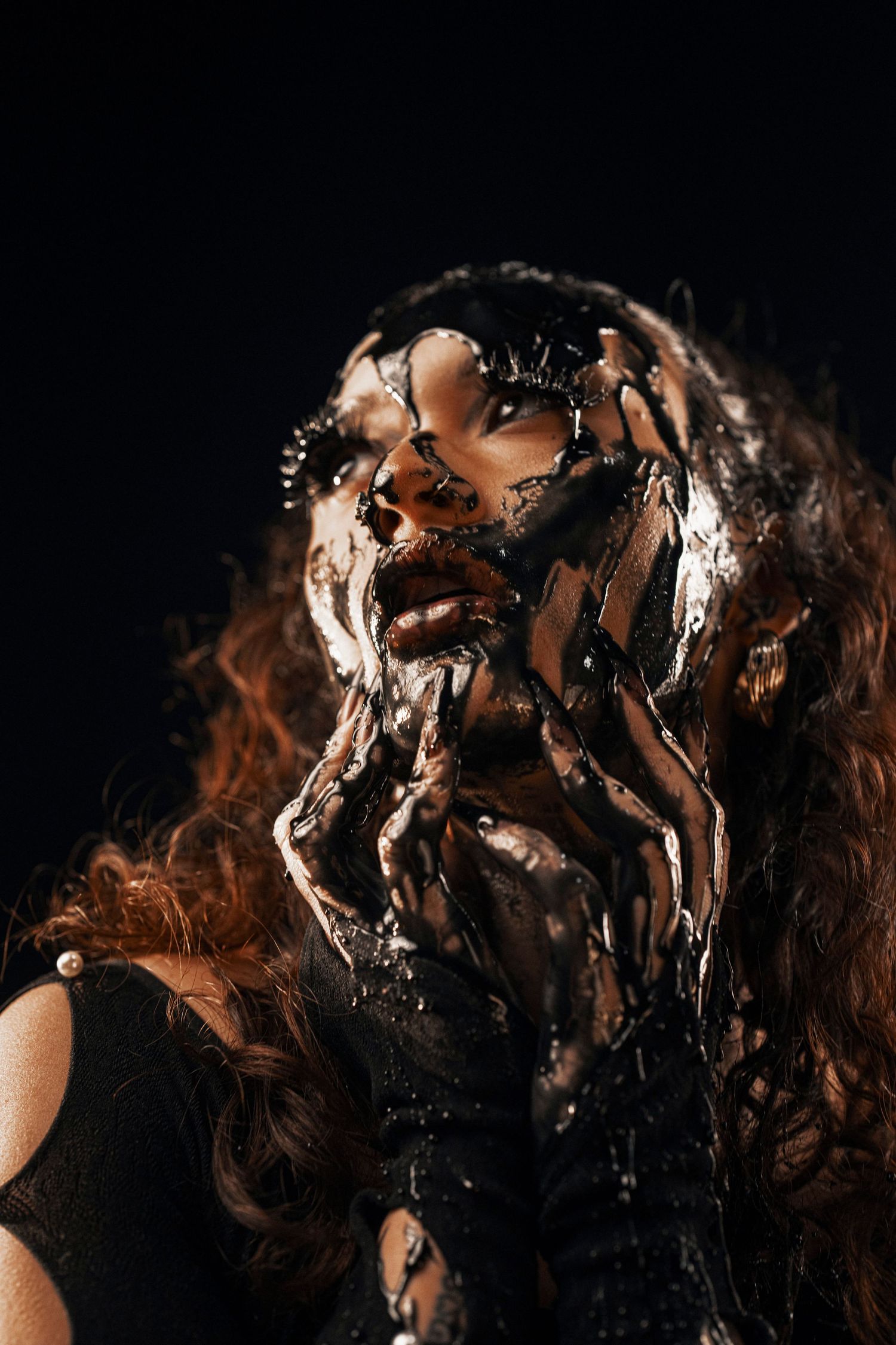 Dark artistic portrait with chocolate syrup dripping and flowing over curly hair against black background.