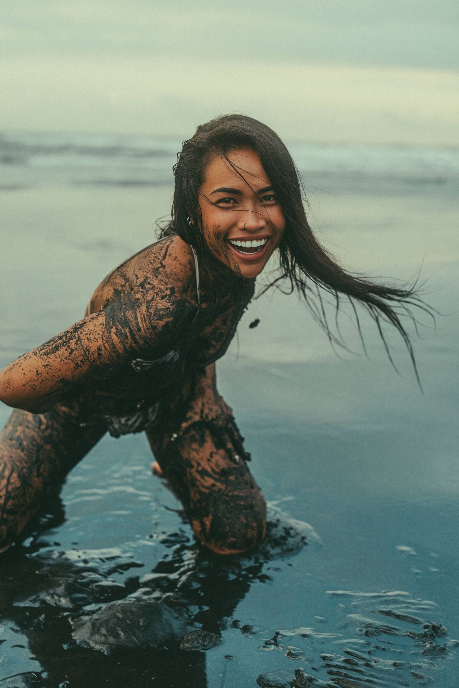 A smiling woman covered in dark mud poses playfully on a beach with ocean waves in the background.