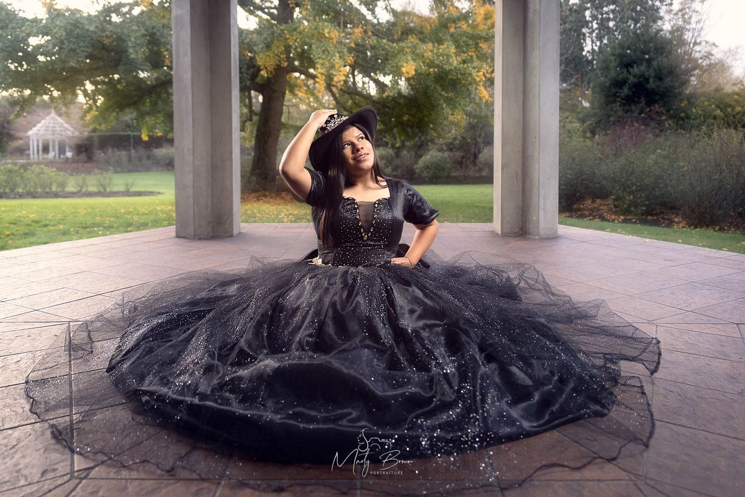 Princess in long black quinceañera flowy dress sitting under baroque arch garden pergola background