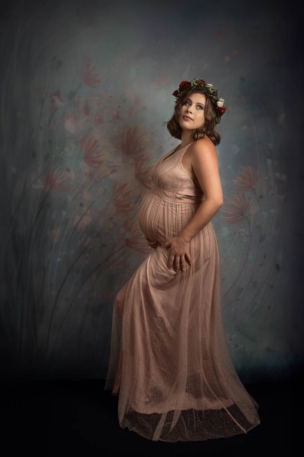 Women in long champagne colour maternity dress with baby bump visible standing side on against floral background
