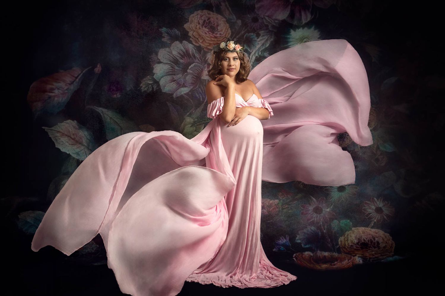 Women in long champagne colour maternity dress with baby bump visible standing material floating all around her against floral background