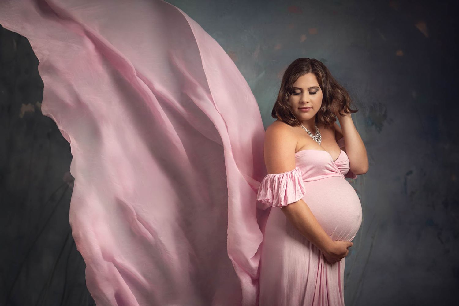 Women in pink maternity dress in studio looking down at her baby bump with floating dress behind her, floral backdrop