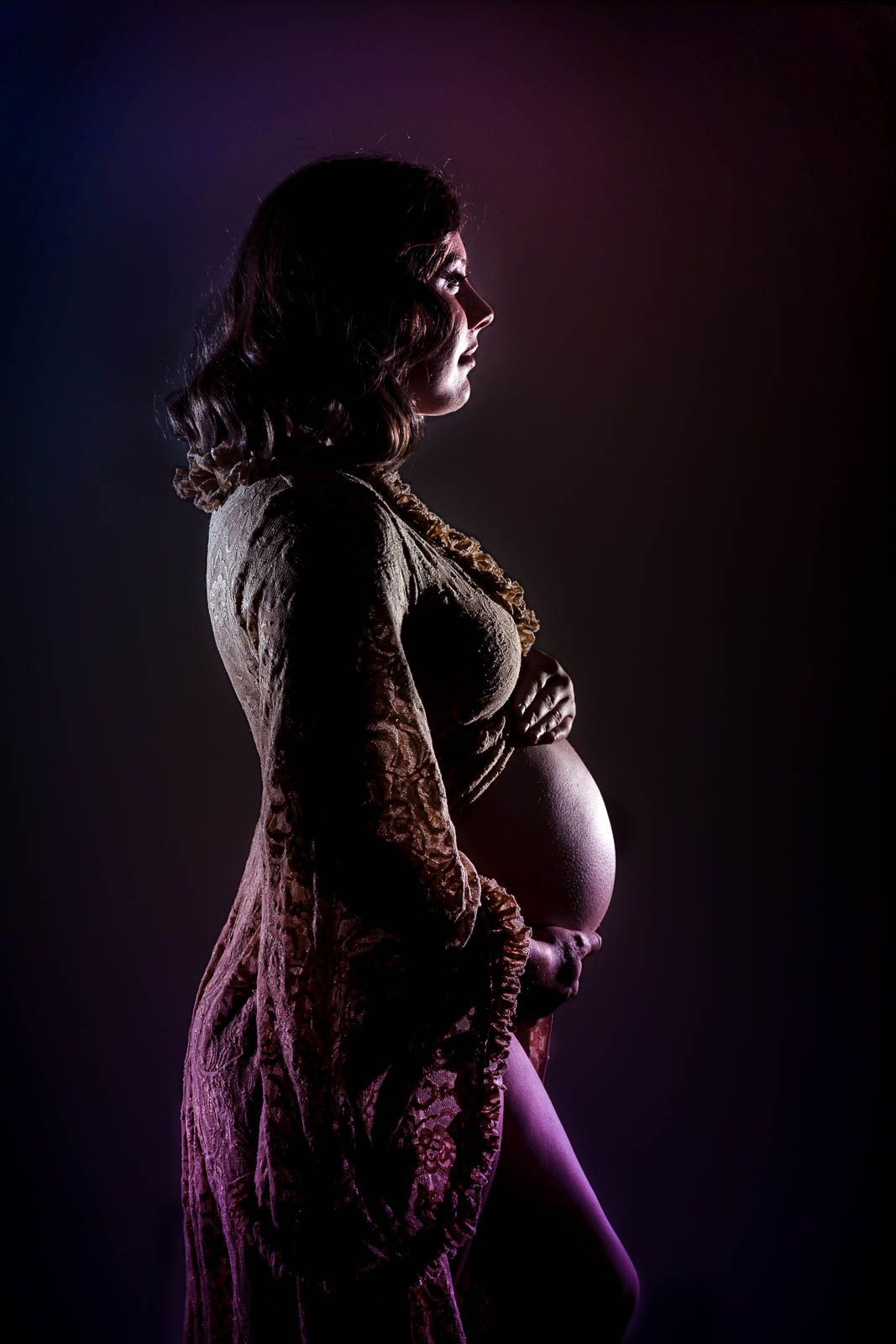 Women in long champagne colour maternity dress with baby bump visible standing side on silhouette against black background