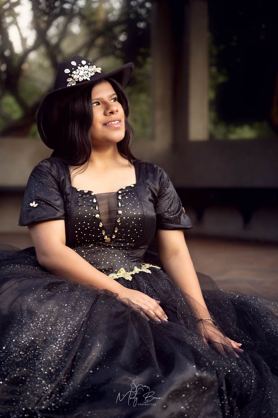 Princess in long black quinceañera flowy dress sitting under garden pergola