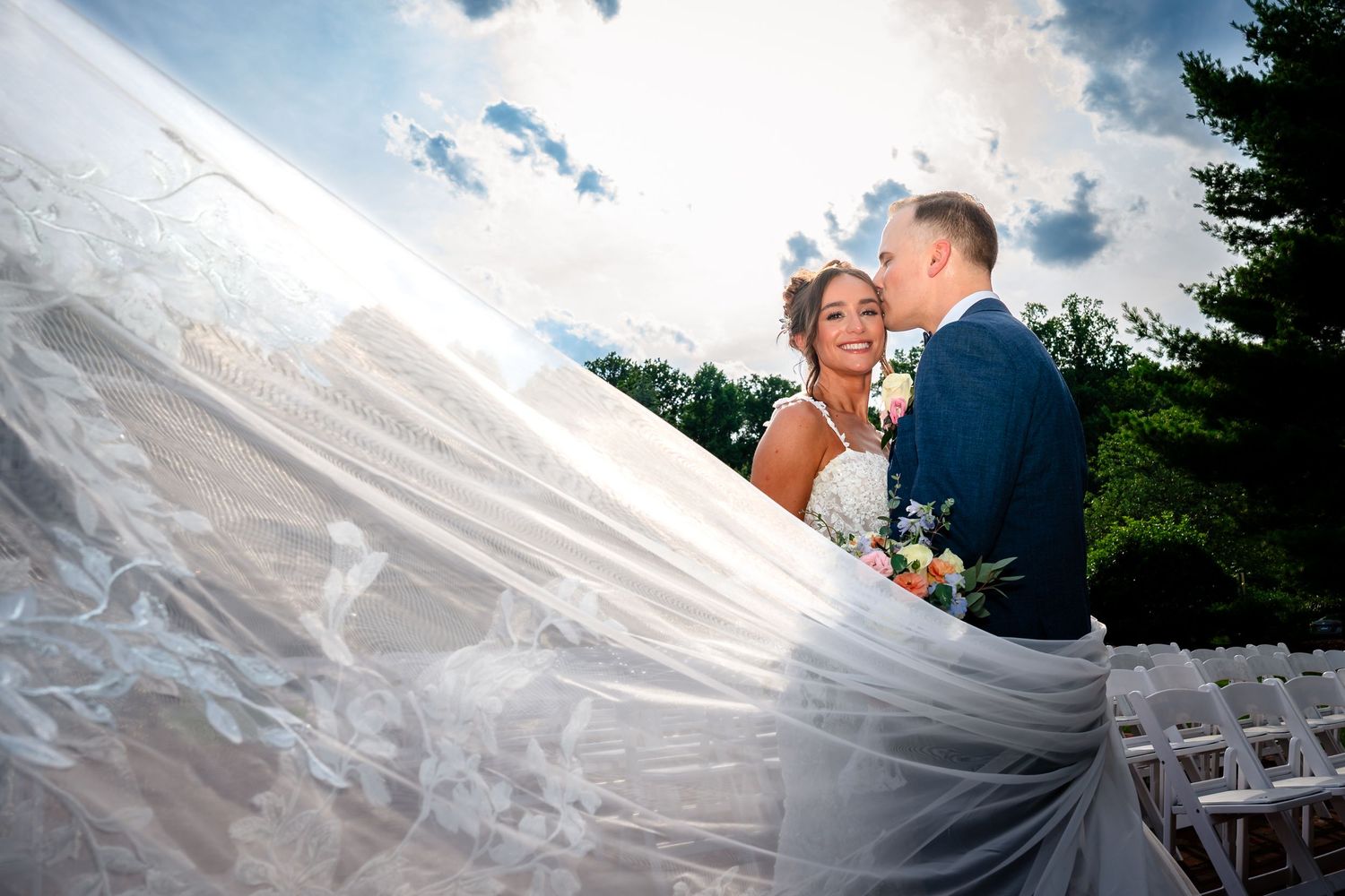 Dramatic wedding portrait with flowing veil against blue sky and evergreen trees.