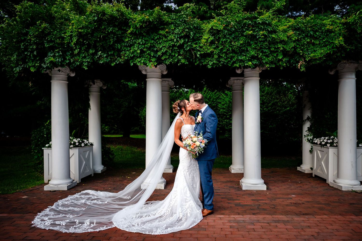 A couple poses between white classical columns along a brick pathway.